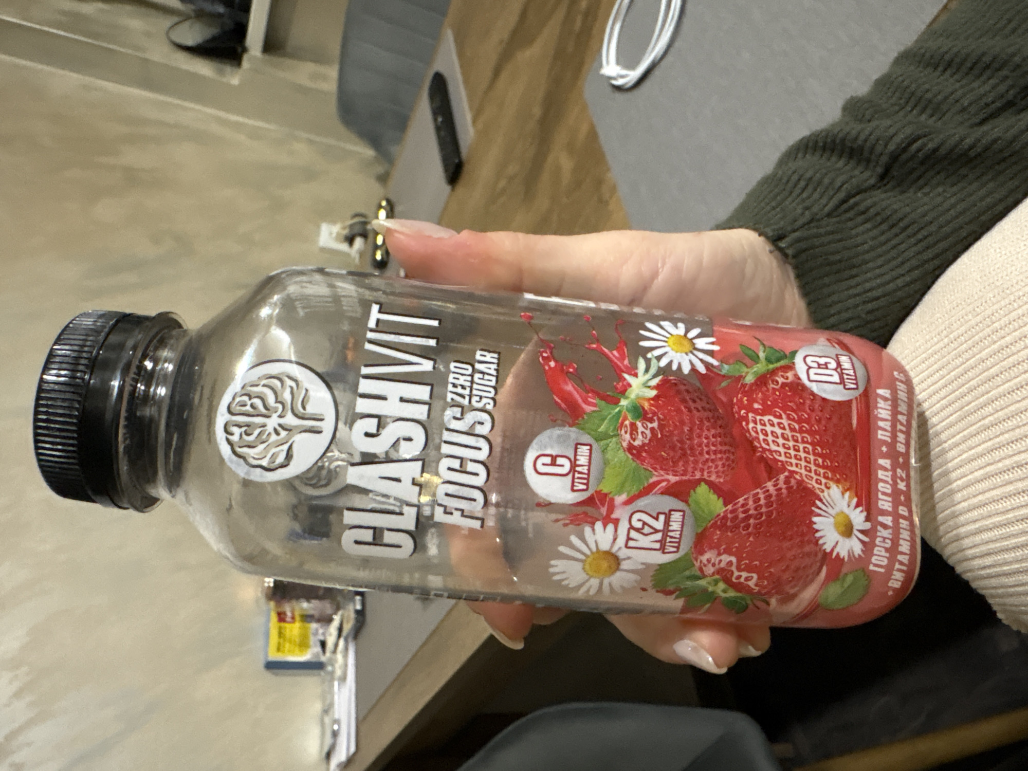 A person is holding a bottle of "Clashvit Focus" zero sugar drink. The bottle is clear with a black cap and has an image of strawberries and daisies on the label. The label also indicates that the drink contains vitamins C, K2, and D3, and includes the text "ГОРСКА ЯГОДА + ЛАЙКА + ВИТАМИН D-K2+ ВИТАМИНС" which translates to "Mountain Berries + Lime + Vitamin D-K2 + Vitamins". The person's hand is visible holding the bottle, and they are wearing a green sweater on one side and a beige sweater on the other. The setting is an indoor environment with furniture in the background. Sofia, Bulgaria is not mentioned in the image and there is no evidence to show that it is located there.