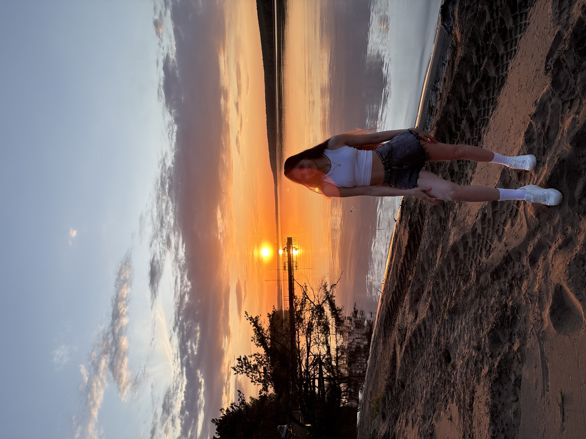 A person is situated on a sandy beach adjacent to a body of water. The setting appears to be at sunset, as indicated by the sun positioned low on the horizon, creating a bright, orange glow. The sky displays a gradient of light blue and orange hues. Some clouds are visible in the sky. In the water, a pier can be seen, and trees are visible along the shore.
It is not possible to confirm the location as Chaykovsky, Russia based on the visual data.