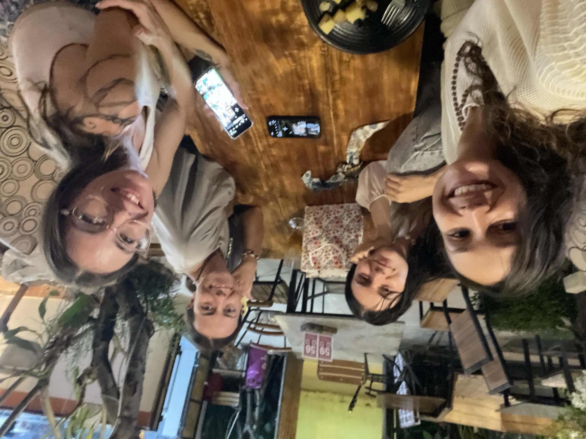 Four individuals are seated around a wooden table at an outdoor cafe, possibly in Phu Thuy Ward, Vietnam, as indicated by the presence of a sign with Vietnamese characters. The individuals are smiling and appear to be interacting, with two holding smartphones. The table is set with a plate of food, silverware, and a drink. In the background, there are more tables and chairs, as well as plants and trees. The lighting suggests it is evening.