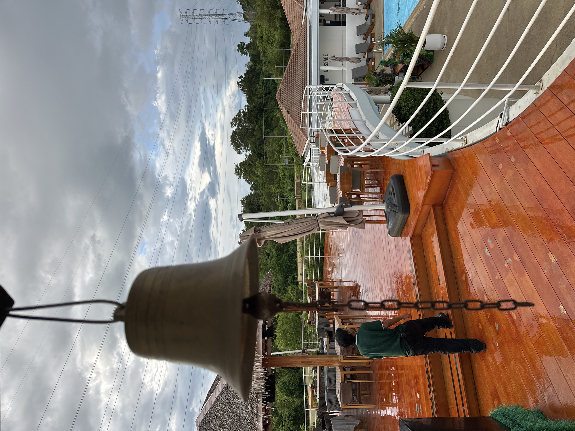 A bell hangs in the foreground of an outdoor dining area with a polished wooden deck, which is wet, indicating recent rain. A person wearing a green shirt and black pants walks up a few steps on the deck. In the background, there are tables and chairs, thatched roofs, a swimming pool, lounge chairs, and lush green trees. Overhead, a power line tower and cloudy sky are visible. The scene appears to be at a resort or restaurant in Kathu, Thailand.