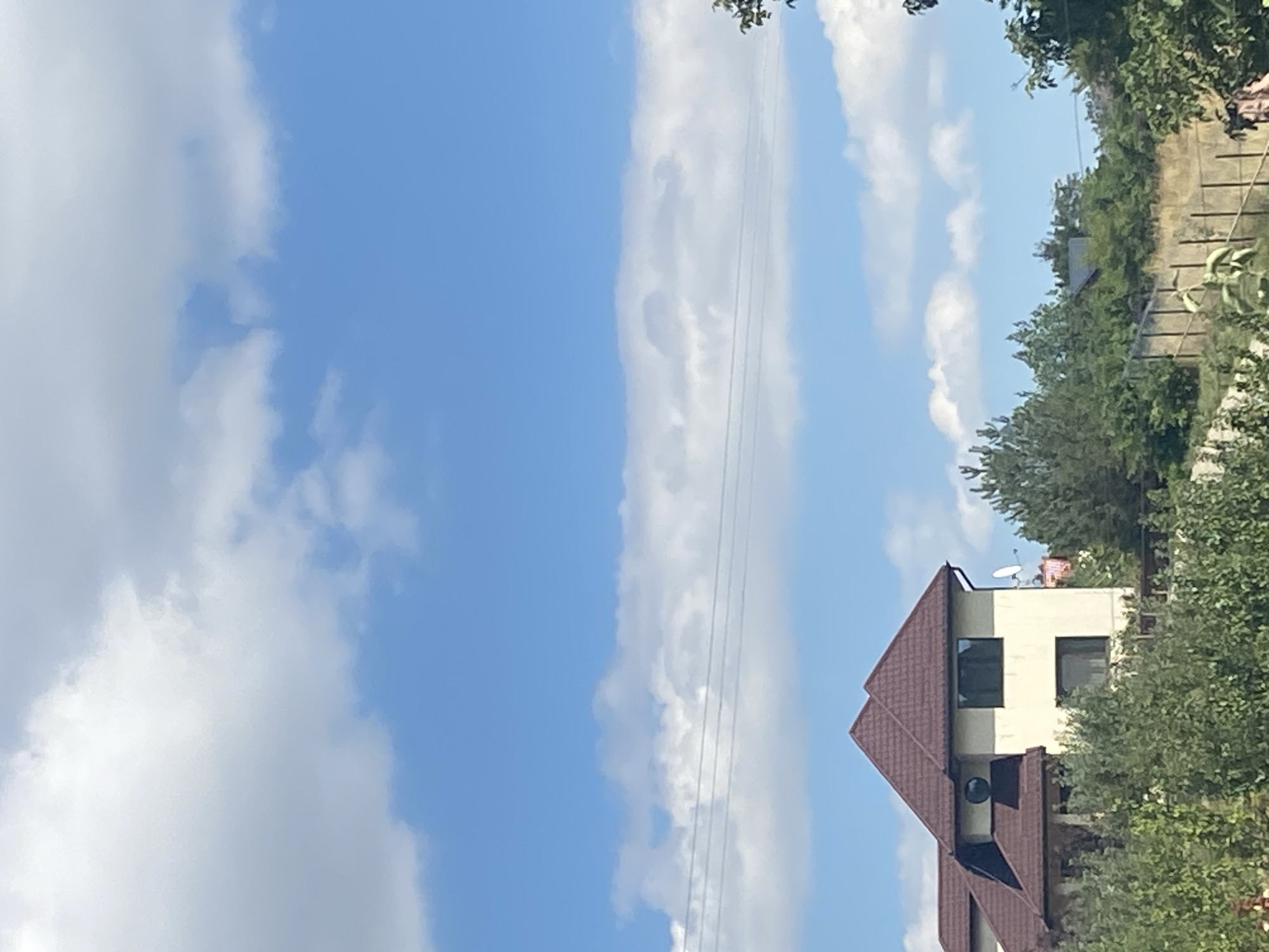 The image shows a residential building with a brown tiled roof, partially obscured by dense green foliage. The building has light-colored walls, dark-framed windows, and a satellite dish on its exterior. Above the building, a bright blue sky is visible, dotted with white, fluffy clouds. In the foreground, the tops of trees and a fence are present. The scene suggests a suburban or rural setting, possibly on a sunny day. The context of Târgu Neamț, Romania, implies this is likely a residential area within or near the city. There are no people clearly visible in the frame.