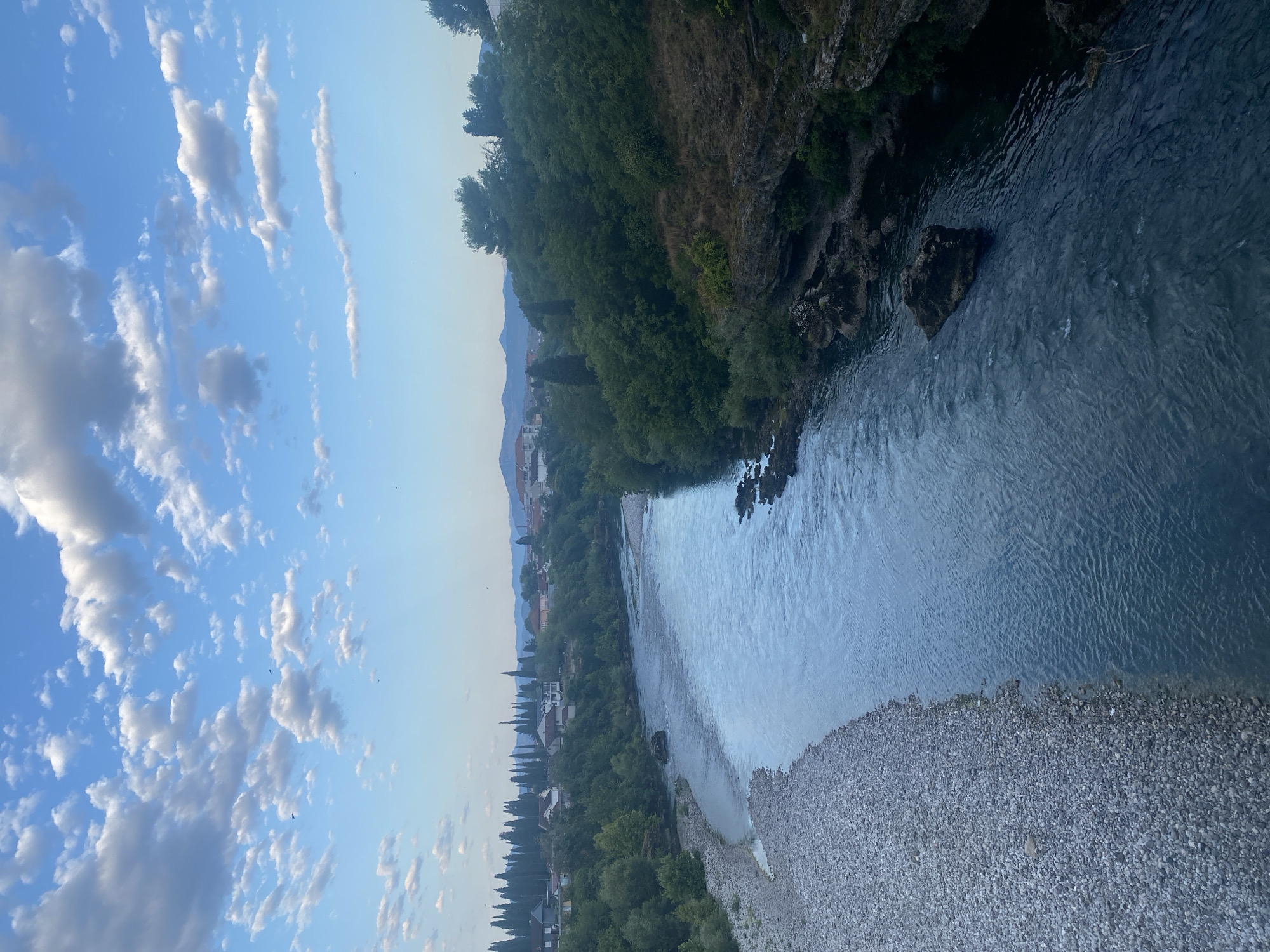 The image depicts a wide river flowing through a city, likely Podgorica, Montenegro, under a partly cloudy blue sky. The river has a stony bank on the left and a rocky, tree-covered embankment on the right. Buildings and cypress trees are visible on the hills in the background, and mountains can be seen in the far distance. The overall impression is of a natural landscape integrated with an urban environment.
