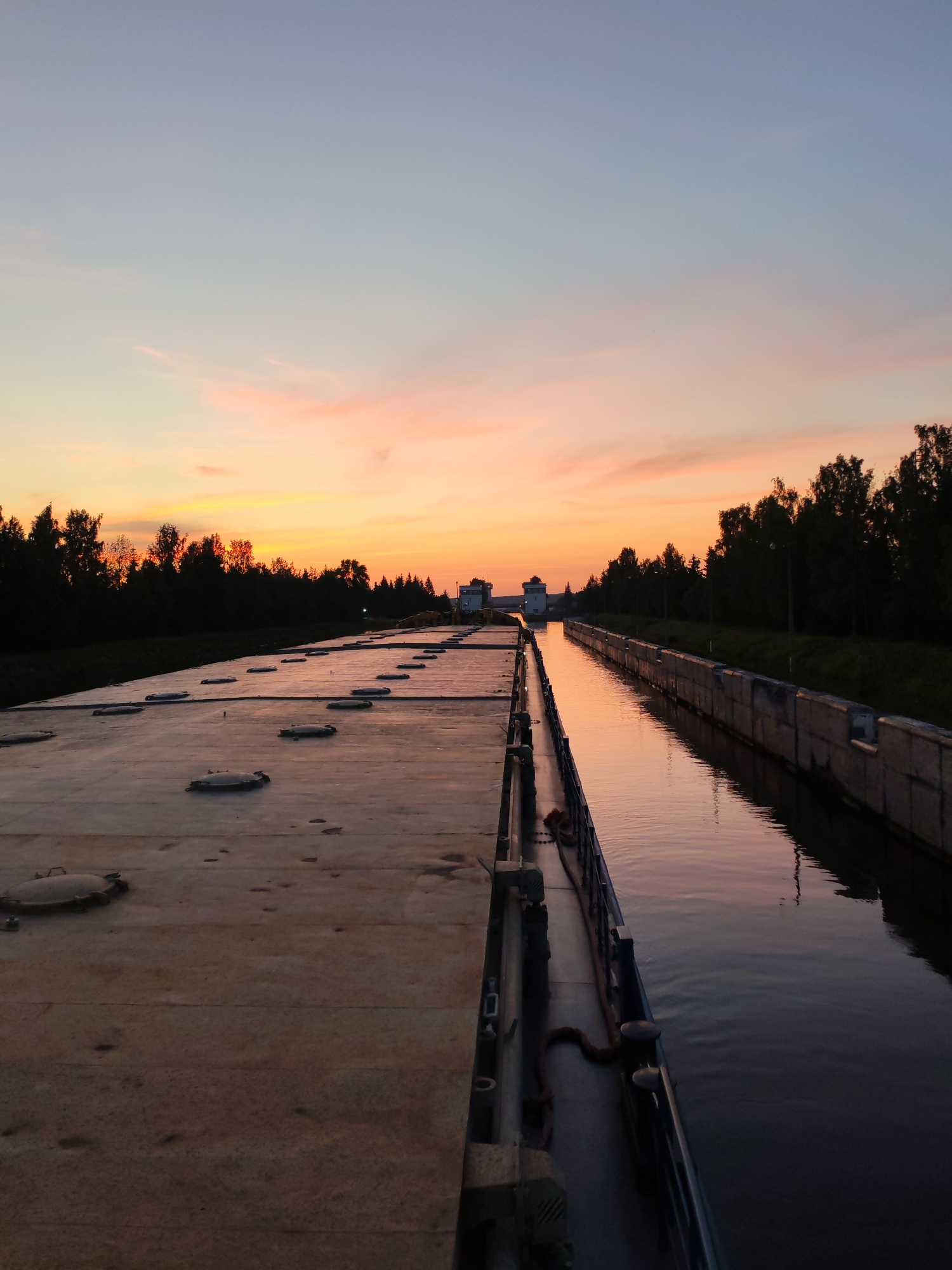 The image captures a sunset over a canal in Vytegra, Russia. The sky is painted with hues of orange, pink, and blue, with wispy clouds. The canal itself is calm, reflecting the colors of the sky. On the left side of the canal, there is a wide, flat structure with several circular hatches. Trees line the right bank of the canal, silhouetted against the colorful sky. In the distance, structures that appear to be part of a lock system are visible. The overall atmosphere is serene and picturesque.