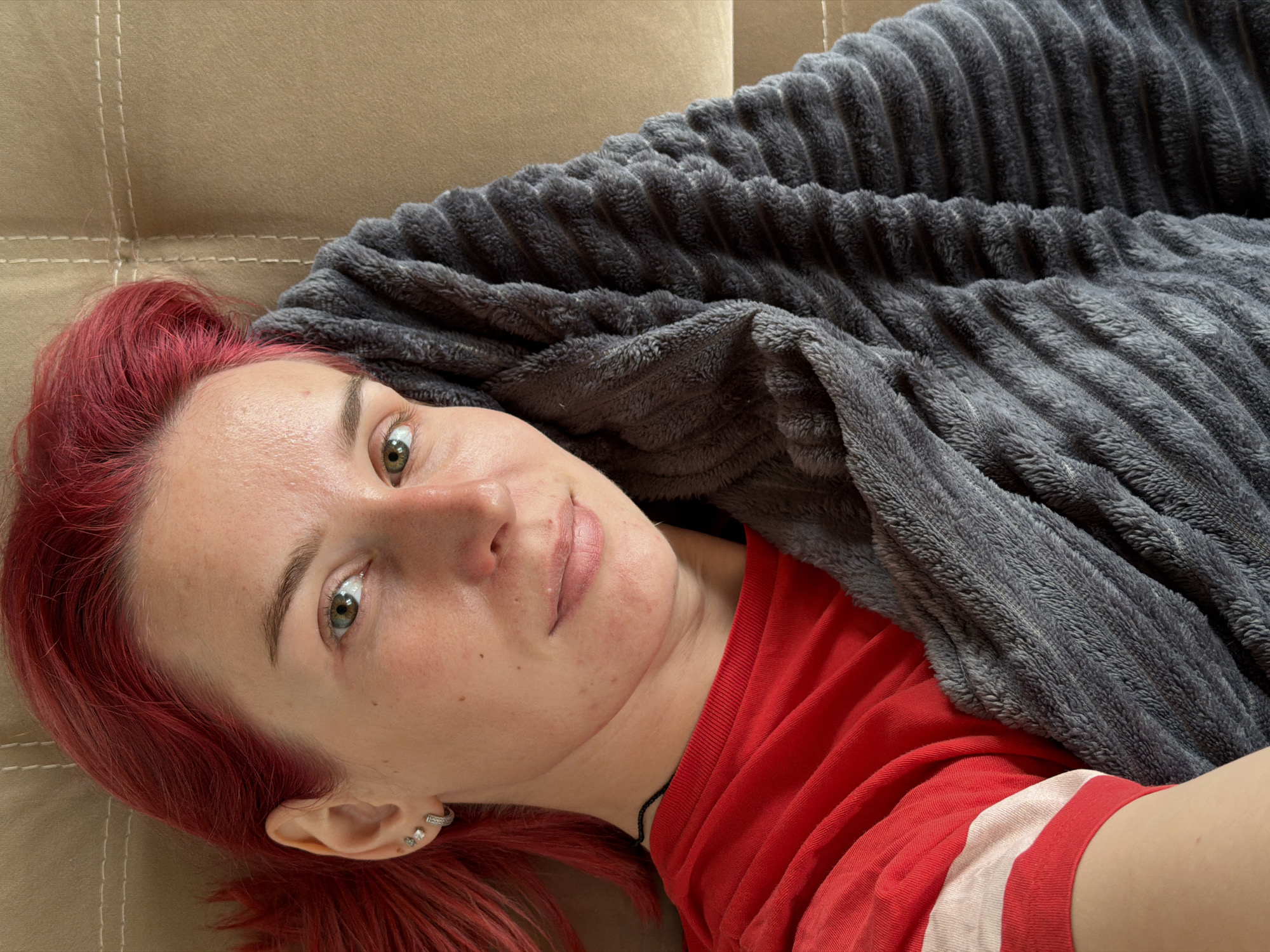 A young woman with vibrant red hair lies on her side, her face turned towards the camera. She is covered by a plush, grey blanket that obscures her body from the chest down. Her left arm is visible, peeking out from under the blanket, and she wears a red t-shirt with a white stripe across the chest. She has a light complexion with a few freckles and small blemishes on her cheeks and forehead. Her eyes are green, and she is wearing small stud earrings in her left ear. A thin black necklace is visible around her neck. The background appears to be a padded, light brown surface, possibly a sofa or a bed. The scene suggests a relaxed, indoor setting. There is no visible indication of the city of Chaikovsky, Russia.