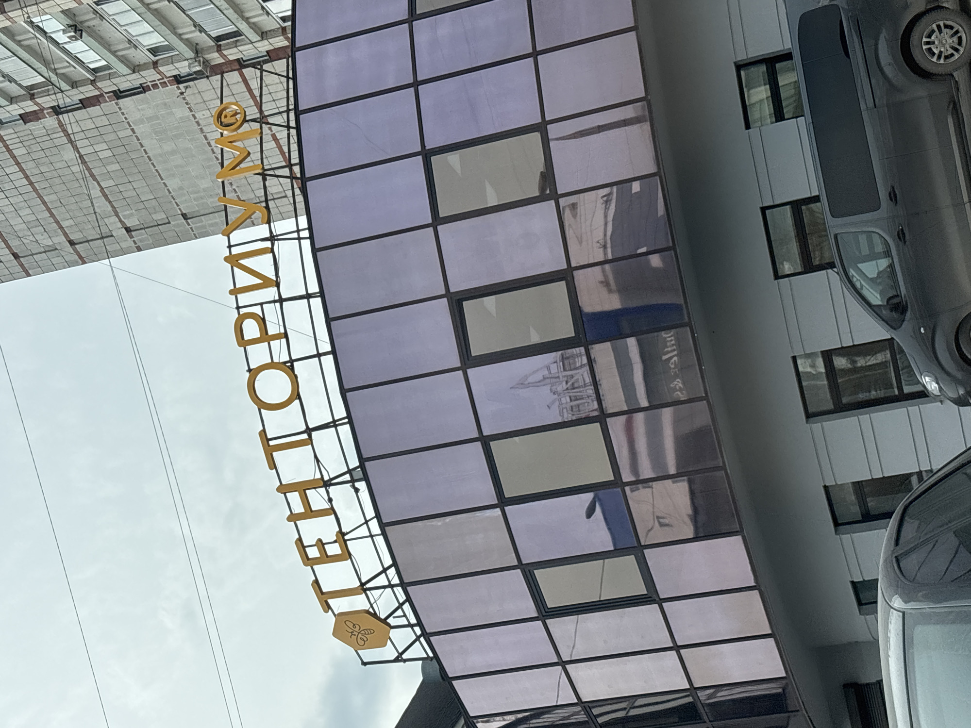 The image depicts the facade of a building in Perm, Russia. A vertical, gold-colored sign spells out "ТЕЛЕНТОРИУМ" (Telentorium) with a hexagonal emblem featuring a bee at the bottom. This sign is attached to a grid-like glass canopy that forms a section of the building's entrance. The reflection in the glass panels shows the architecture of an adjacent building. To the right of the building are parked vehicles, appearing to be vans, partially obscuring the facade. The sky is overcast and partially visible in the upper left corner. The building appears to be situated in an urban area.