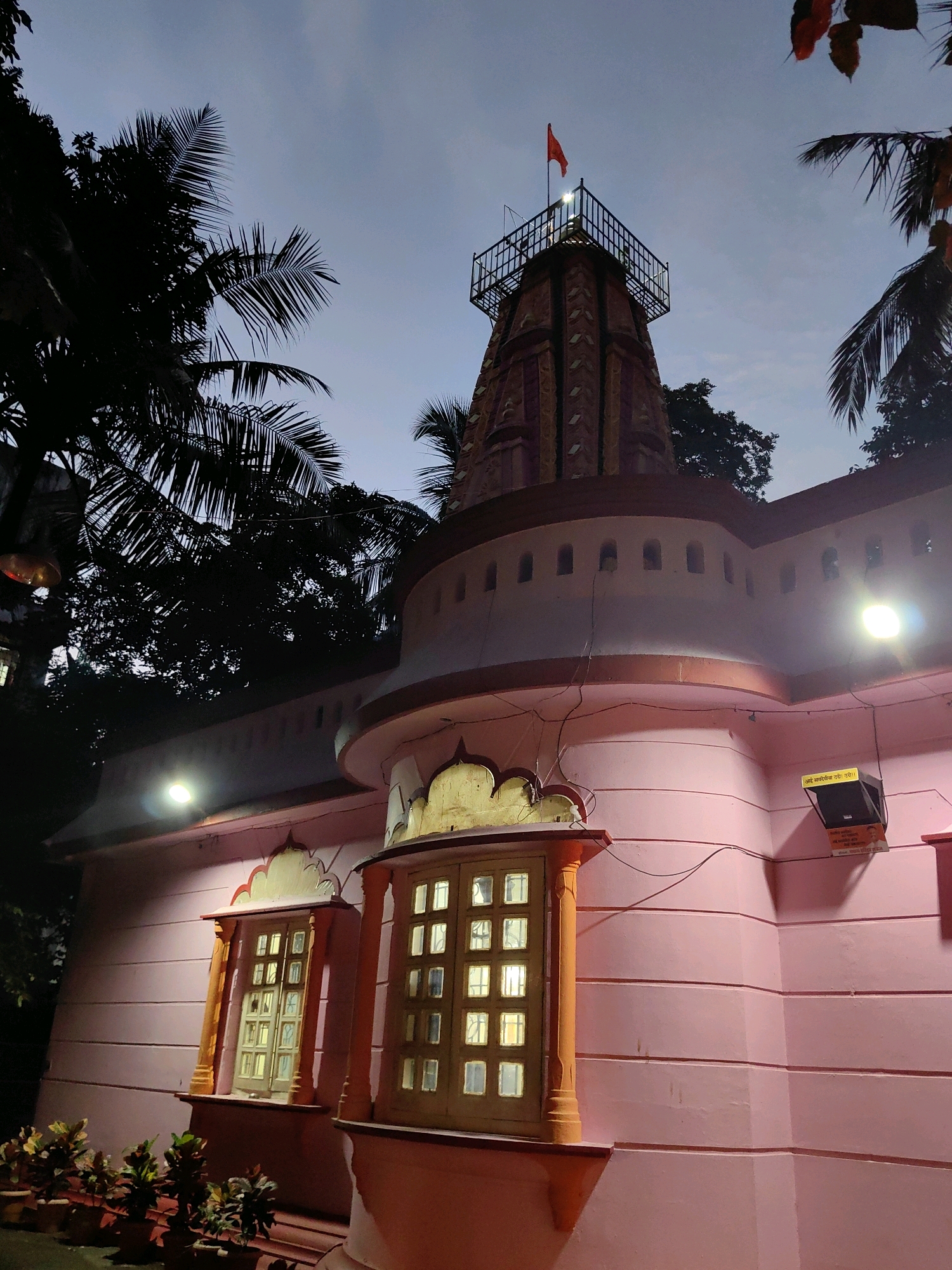 The image depicts the exterior of a temple in Mumbai, India, at dusk. The temple has a light pink facade with horizontal grooving. Two ornate windows with multiple panes, illuminated from within, are visible on the lower section of the building. Above these windows, a rounded architectural feature runs along the temple's circumference, punctuated by small, arched openings. To the left of the rounded section, palm trees with dark foliage are silhouetted against the sky.
The uppermost part of the temple features a tower, possibly a Shikara, constructed with reddish-brown stone-like blocks. A black metal railing surrounds the top of the tower, and a small orange flag is visible atop it, fluttering in the breeze. Lights are installed on the temple's exterior, illuminating the facade and some of the architectural details. A security camera and a sign with text in an Indian script are mounted on the upper right side of the building. Potted plants are arranged along the base of the temple on the left side. The sky is a dusky blue, with soft lighting conditions.