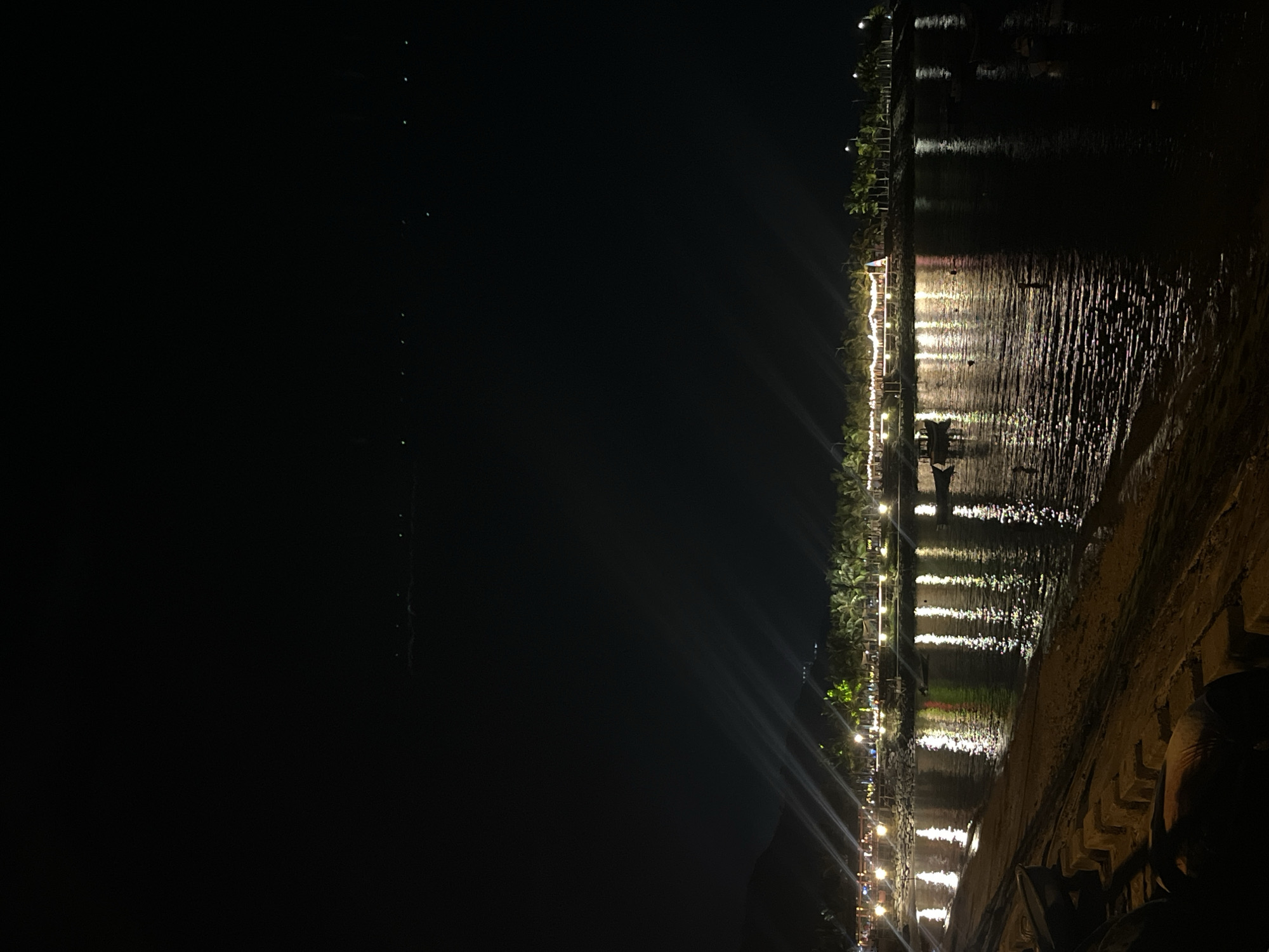This photograph captures a nighttime scene along the coast of Nha Trang, Vietnam. The foreground features a dark sandy beach with a stone embankment constructed from large concrete blocks. The water of the sea is visible, reflecting the warm glow of lights from the distant shore.
The middle ground is dominated by a vibrant coastline illuminated by numerous lights. Buildings and structures are lined with string lights and individual lamps, creating a festive and inviting atmosphere. Palm trees are interspersed along the shore, their silhouettes softened by the artificial lighting. A narrow strip of beach is visible between the water and the illuminated structures. Several small boats are moored in the water, with one boat appearing to be in motion with a few individuals onboard.
The background consists of a dark, starless night sky, with distant lights faintly visible on the horizon, suggesting the presence of a more distant settlement or activity. A dark, mountainous landscape is visible to the far left of the image, partially illuminated by some of the coastal lights. The overall impression is a lively and picturesque beachfront at night, likely a popular tourist area in Nha Trang.