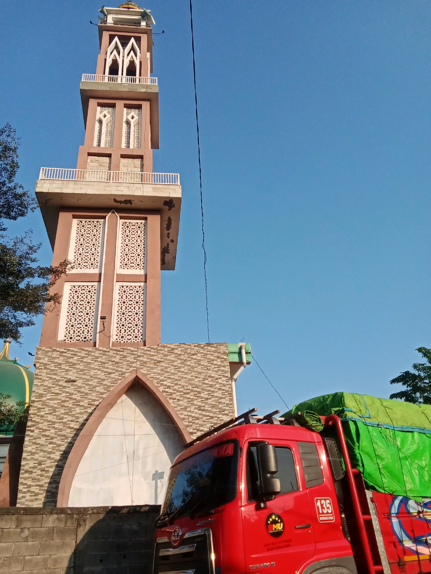 The image shows a bright, clear day in Keboharan, Indonesia, with a tall mosque minaret dominating the skyline. The minaret is a striking tower with multiple levels, featuring decorative windows with intricate patterns. The lower section of the minaret is clad in stone tiles. Adjacent to the minaret, a portion of the mosque's green dome is visible. In the foreground, a red truck is parked, its bed covered with a bright green tarp. The truck has various markings, including the number "135" and a company logo. A low brick wall runs along the bottom of the image, casting a shadow. The clear blue sky suggests it is daytime, and the overall scene depicts a typical street view in the area.