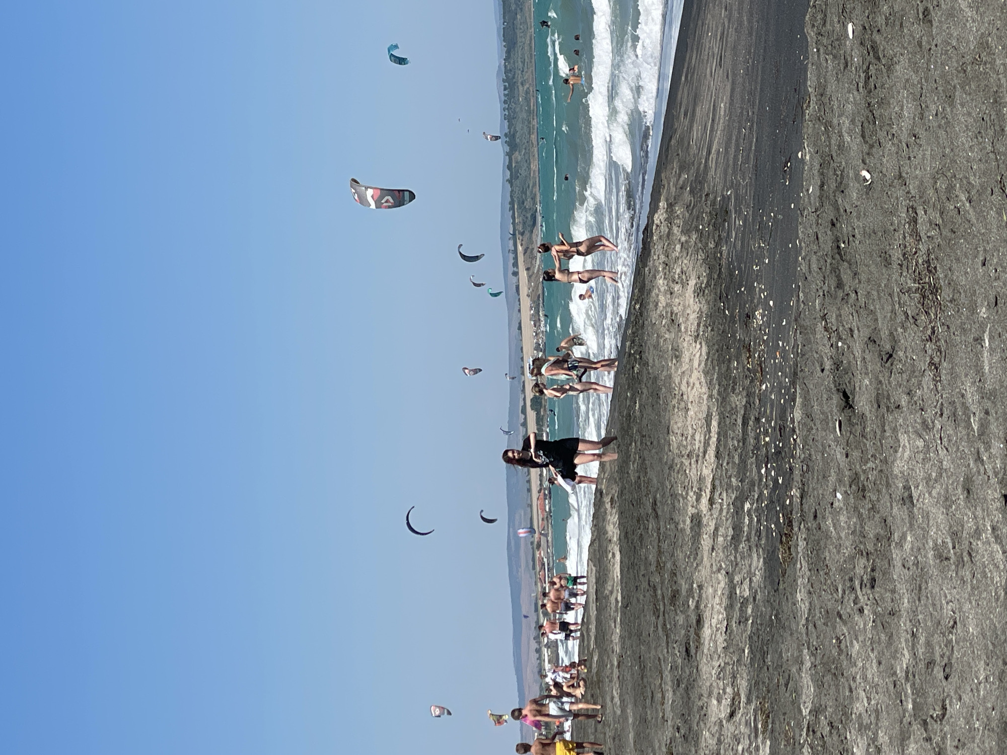 A sunny day at the beach in Burgas, Bulgaria, where a variety of people are enjoying the water and the sand. The scene is dominated by a wide, dark sand beach that meets the turquoise waters of the sea. Numerous kites are visible in the sky, indicating a popular spot for kitesurfing. Several individuals are in the water, some swimming and others appearing to be engaged in water sports. On the beach, groups of people are scattered, some walking, some sitting, and some standing near the shoreline. The sky is clear and blue, with a distant, hazy coastline visible in the background.