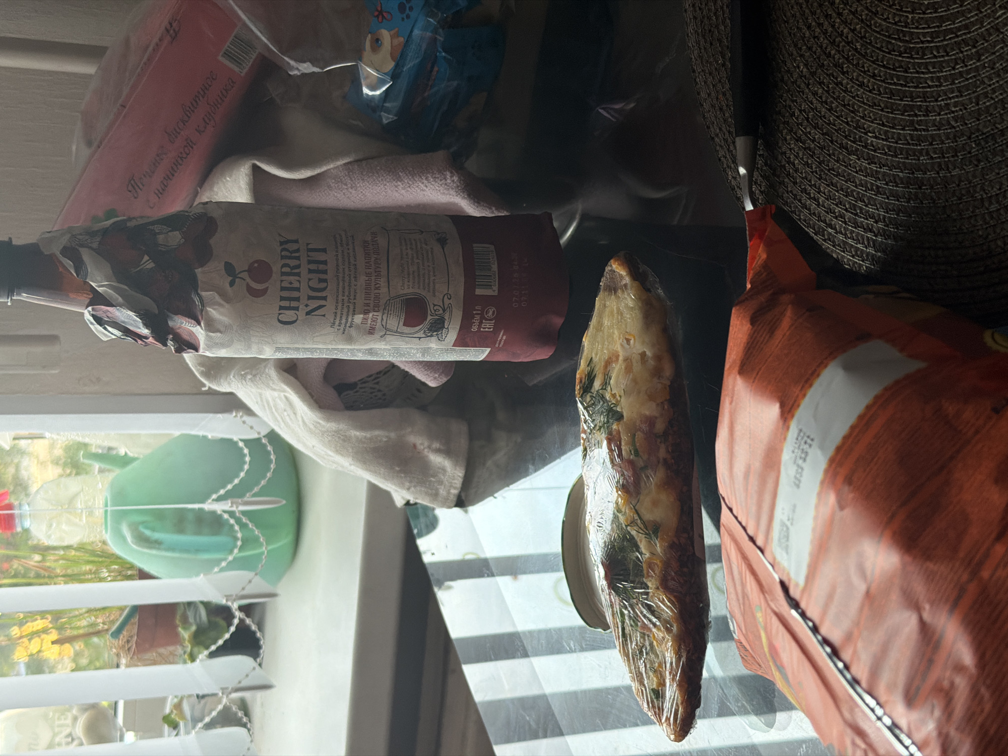 The image depicts a close-up view of a table inside a residential setting, likely a kitchen or dining area, located in Kolpino, Russia. The table's surface is reflective and shows parallel lines, possibly from a placemat or a patterned tablecloth.
Dominating the foreground is a partially visible orange package, likely containing snacks, positioned diagonally across the table. To its left, a piece of food, possibly a slice of pizza or focaccia, is wrapped in transparent plastic.
In the mid-ground, a bottle with a label reading "CHERRY NIGHT" stands prominently. The bottle is wrapped in a decorative paper featuring cherries and text in Russian. Behind the bottle, a red box with Russian text, possibly a confectionery item, is visible. A white cloth is draped over some of the items.
In the background, a window with white vertical blinds allows natural light to illuminate the scene. Outside the window, a green watering can is visible, along with potted plants and a water bottle.
The overall impression is of a casual, domestic scene, with food and beverages present on a table. No people are visible in the image.
