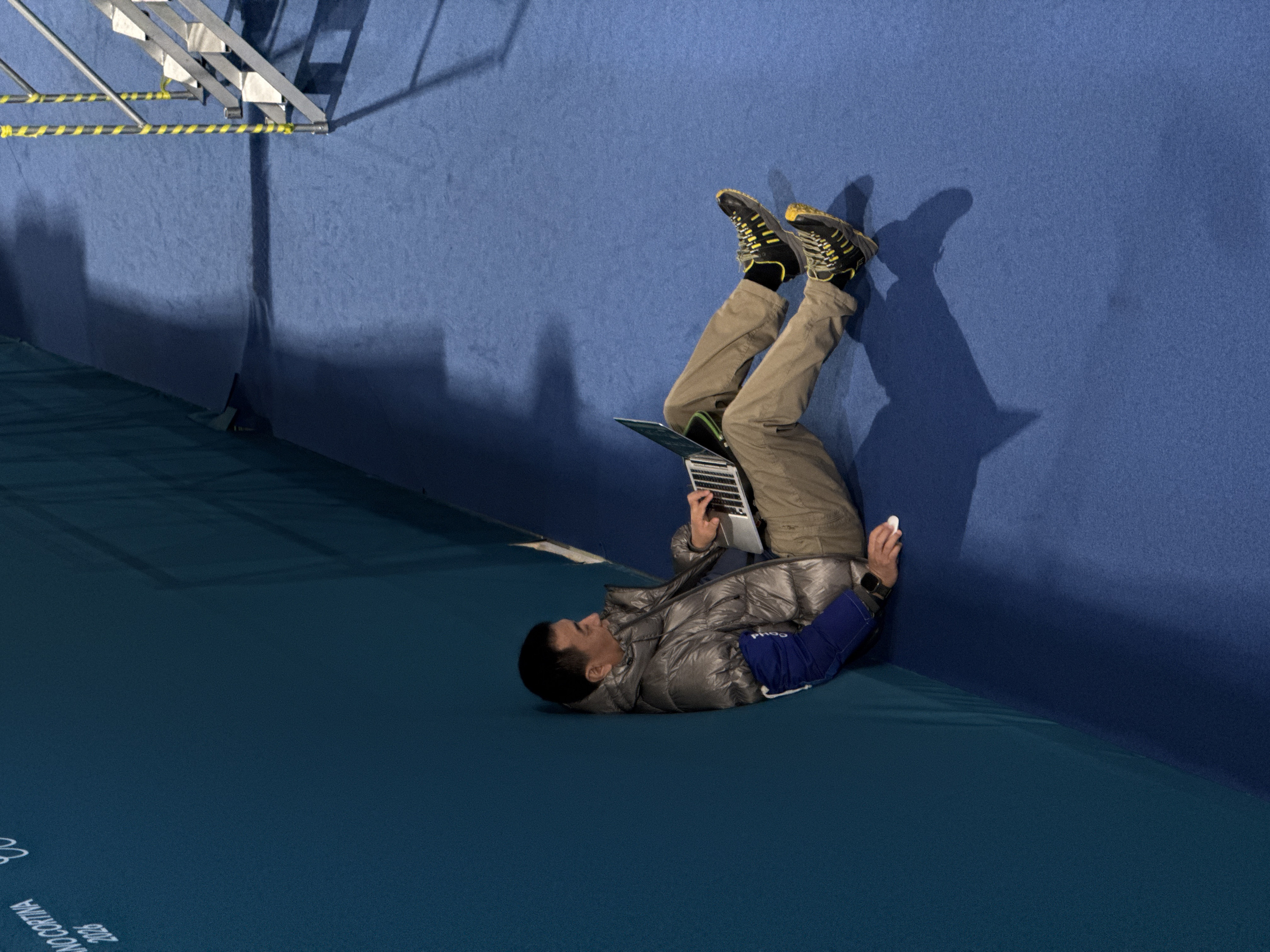 The image captures a unique, almost gravity-defying scene featuring a person lying on their back on a teal-colored floor, with their legs propped up against a dark blue wall. The person, appearing to be an adult male, is dressed in practical attire suitable for cooler weather or an active environment. He wears a shiny, silver-grey puffer jacket with blue long sleeves, khaki or tan-colored cargo pants, and black and yellow athletic shoes with visible laces. Black socks are also visible between his pants and shoes.
In his hands, he is actively engaged with a silver laptop, which rests on his stomach or chest, angled so he can type on its keyboard. His fingers are positioned over the keys, suggesting he is working or browsing. In his right hand, he holds a small, white, seemingly wireless earbud or similar small electronic device. A black smartwatch is visible on his left wrist.
The floor is a flat, matte teal surface, while the wall is a darker, matte blue. Both surfaces appear to be soft or padded, possibly indicating a sports facility, gym, or a temporary event space. On the floor, near the bottom left corner, there is faint white text that reads "MILANO CORTINA 2026", hinting at the location or event affiliation.
In the upper left corner of the image, a metallic structure is visible, composed of silver-colored beams or scaffolding. A horizontal bar within this structure is wrapped with yellow and black striped tape, commonly used for hazard warnings or demarcation. Strong shadows are cast across the blue wall and the floor, originating from the person and the metal structure, suggesting a prominent light source from the upper right or left. The overall lighting is dim but focused, highlighting the central figure. The pose of the person, lying down with legs up the wall, suggests a moment of comfortable rest or an unconventional working posture.
