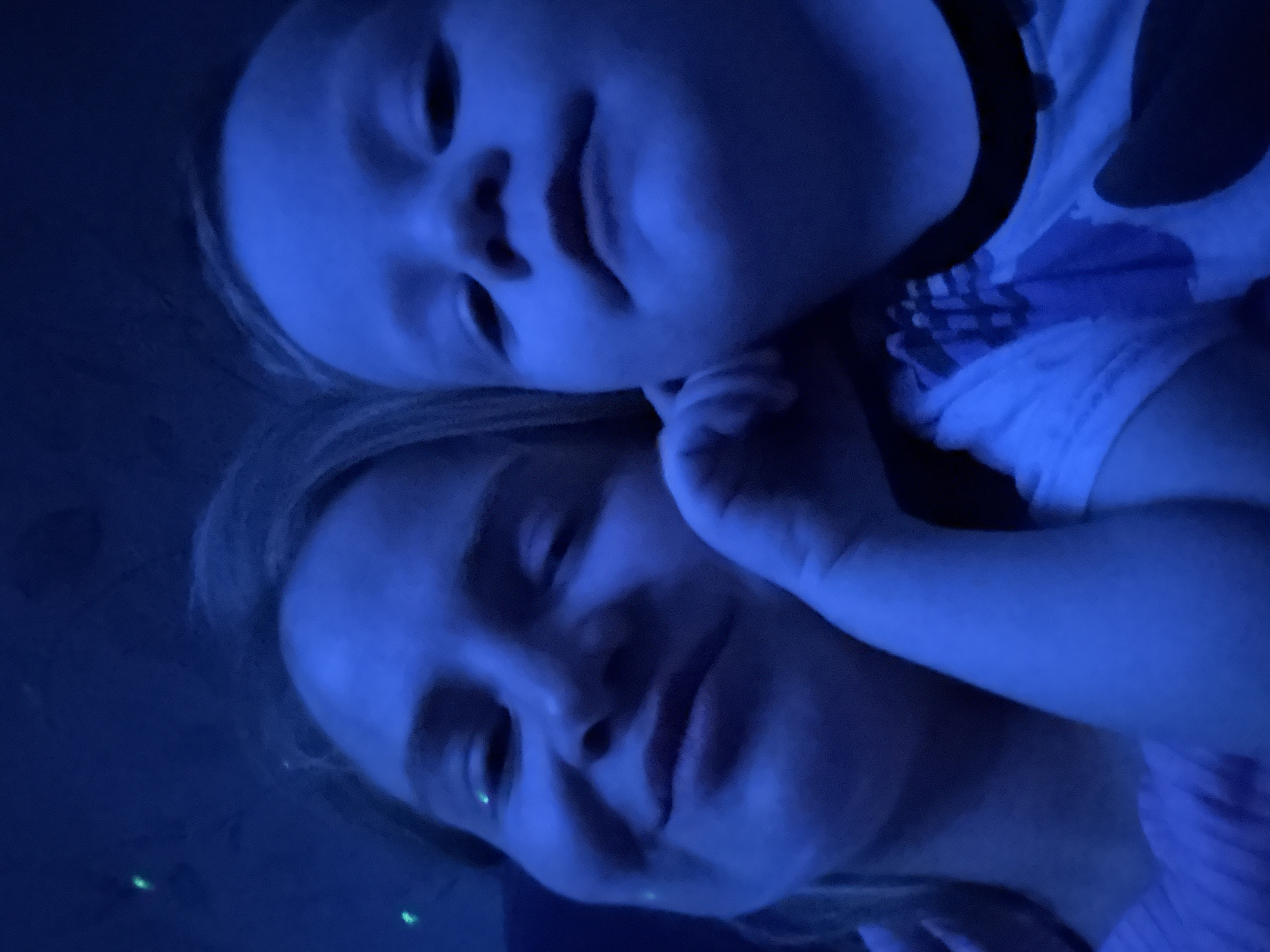 A selfie-style photograph shows an adult female and a young child together in a blue-toned environment. The child is positioned above and slightly behind the woman, with their faces close together. The woman appears to have her eyes closed and is smiling softly. The child has a slight smile as well. The photo is taken at an angle, suggesting a handheld camera. The presence of this scene in Saint Petersburg, Russia cannot be determined without additional information.
