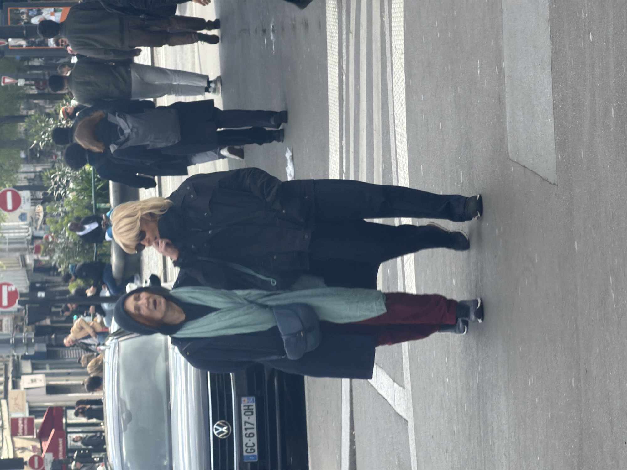The image captures a street scene in Paris, France. Focus is on two women walking. The first woman has blonde hair and wears sunglasses, a black jacket, and black pants. The second woman, closer to the camera, has brown hair, wears a black hooded coat and a light green scarf. A navy blue purse is slung across her body. They stand near a black Volkswagen car. The car's license plate reads "GC-617-QH-92." A crosswalk is visible on the street, and blurred figures of other pedestrians populate the background. Street signs and buildings are visible in the background, suggesting a busy urban area.