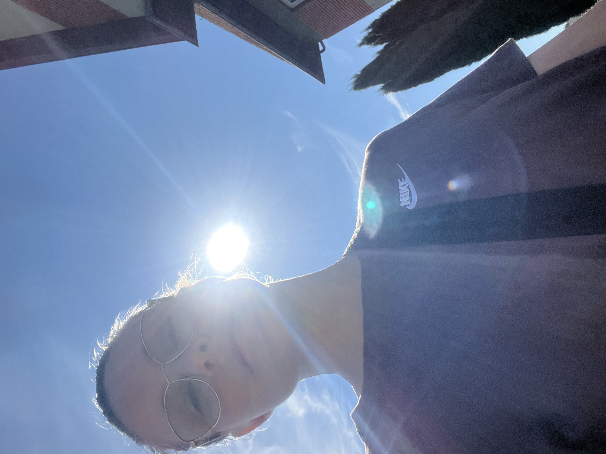 A low-angle, outdoor selfie shows a person wearing glasses and a dark-colored t-shirt with a Nike logo. The sun is high in the sky and is causing a lens flare that obscures part of the person's face. In the background, there is a brick building and several tall, dark green trees. The sky is bright blue with a few wispy clouds. There is no information in the image to suggest a location in Saint-Vith, Belgium.