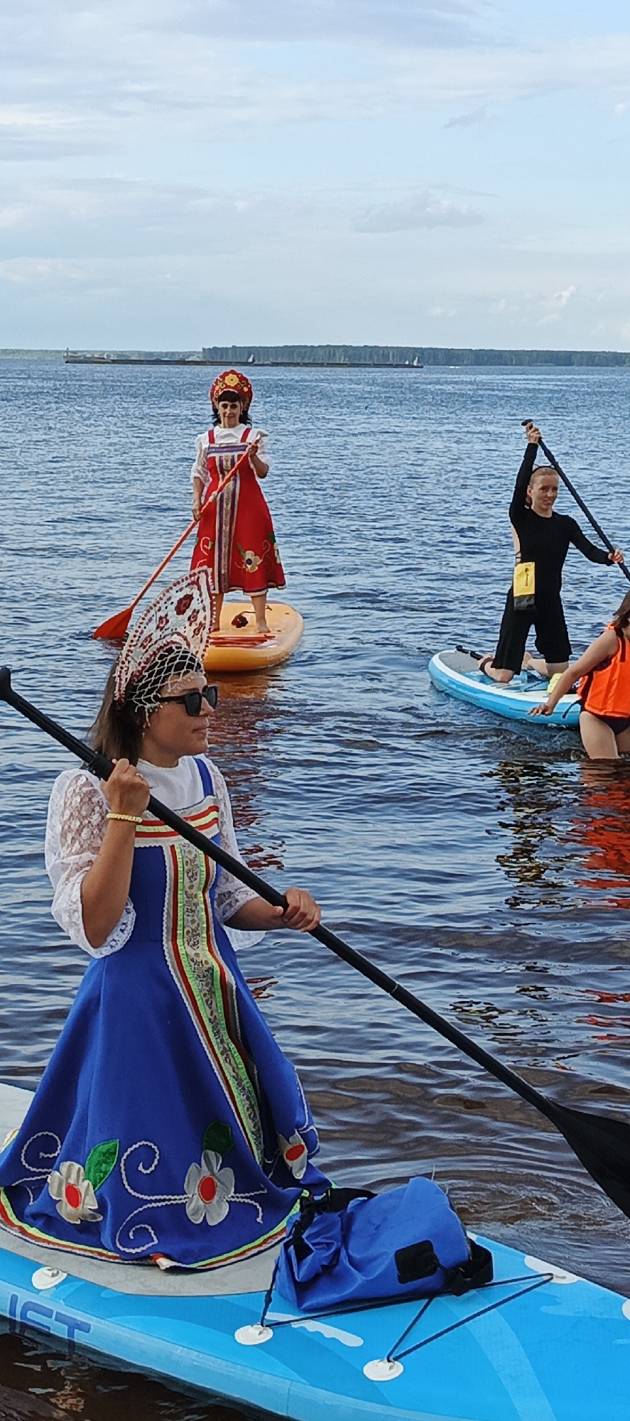 In the city of Chaikovsky, Russia, four women are stand-up paddleboarding on a large body of water, possibly the Kama River, under a partly cloudy sky. The water is calm and deep blue. In the foreground, a woman wearing a blue dress adorned with embroidered flowers and a decorative headpiece with white netting and beads is holding a paddle. She is wearing sunglasses and a yellow bracelet. Behind her and to the left, another woman in a traditional red dress with intricate embroidery and a red headdress stands on a yellow paddleboard, holding a paddle. Further back and to the right, a woman in a black long-sleeved outfit kneels on a blue paddleboard, holding a paddle aloft. A fourth woman, partially visible in the background to the right, is wearing an orange life jacket and is also on a paddleboard. The coastline is visible in the distance with a line of trees.