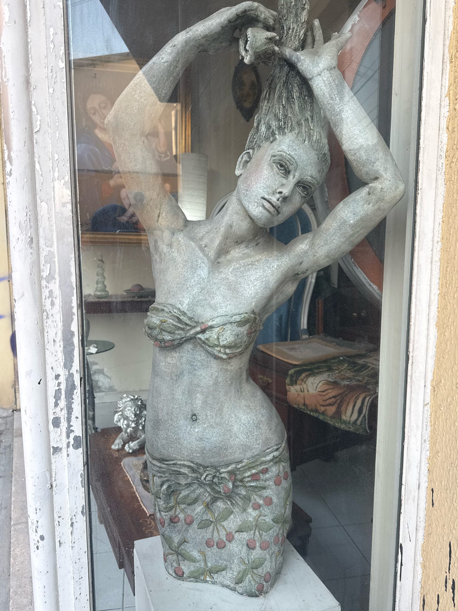 The image shows a statue of a woman with her arms raised, holding her hair. She is wearing a bikini top and a skirt decorated with cherries. The statue is made of a gray material and appears to be in a shop window. Behind the statue, a reflection shows a painting of a woman with glasses and another painting of a face. The shop window is framed by a peeling white painted wooden structure. To the right of the frame, a yellow stucco wall is visible. Inside the shop, a tiger-striped fabric is visible on a piece of furniture. A small metal lion statue sits on a dark wooden surface. The environment suggests an indoor display, possibly a retail space or a gallery, located in Limassol, Cyprus, as indicated by the presence of the statue and its context within a shop.