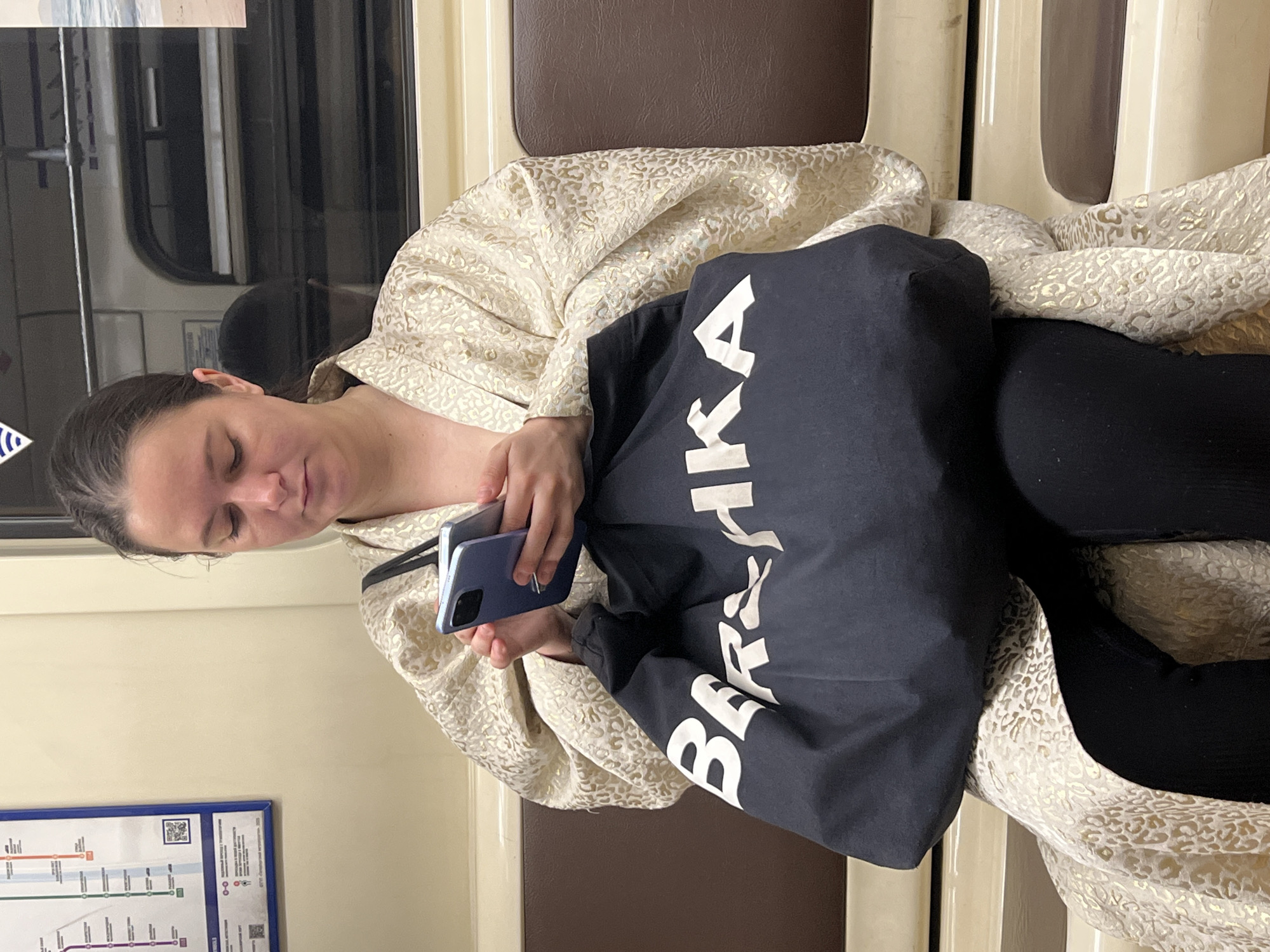 In the interior of a train carriage, a fair-skinned woman with dark hair sits using a blue smartphone. She is wearing black leggings and a light-colored robe with a gold leopard print. A black bag with white lettering is positioned in her lap. The scene is likely within Saint Petersburg, Russia, given the presence of a train map with Cyrillic text on the wall behind her. The train has seats with brown and beige upholstery, and a window with a partial view of the exterior is visible.