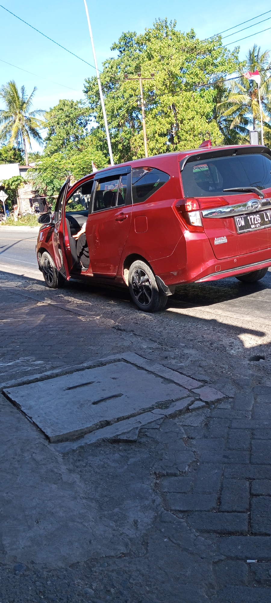 A red sedan is parked on the side of a street in Pakkanna, Indonesia. The driver's side door is open, and a person is seated in the driver's seat, their arm resting on the open door. The car is a Toyota Avanza, and its license plate reads "DW 1726 LY". The street appears to be made of concrete and asphalt, with a drainage grate in the foreground. Trees and buildings line the street, and the sky is clear blue. A flag, possibly the Indonesian flag, is visible in the background. The scene is lit by bright sunlight, casting shadows on the ground.