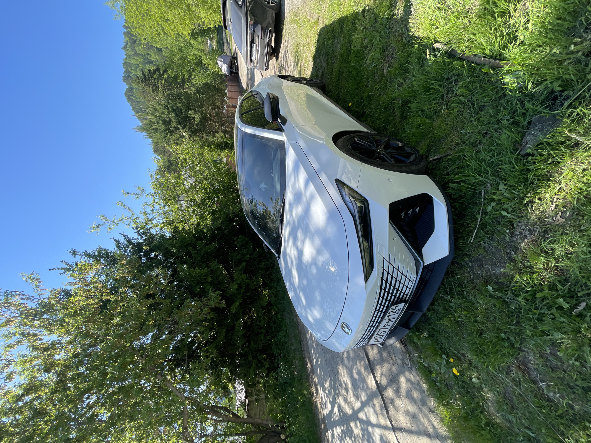 The image displays a white Changan UNI-V sedan, identifiable by its distinctive front grille and logo, parked on a patch of vibrant green grass adjacent to an unpaved dirt road. The car bears a Russian license plate, M057PM 124, indicating registration in Krasnoyarsk Krai. Strong sunlight casts dappled shadows from overhead trees across the vehicle's hood and the road surface, suggesting a clear day.
The immediate environment features dense green grass and various tree species, including deciduous trees with light green foliage and darker coniferous trees. To the left, a rustic wooden fence is partially visible through the foliage. The dirt road slopes uphill, leading to other objects in the background. Further up the incline, a dark-colored sedan or wagon (possibly another Changan vehicle) is parked on the right, with partial license plate information "007" visible. Between the white car and the dark car, a grey tarp-covered object, potentially a utility vehicle or trailer, is parked off the road.
The background is dominated by a substantial, tree-covered hill, densely forested with various shades of green. Above the hill, the sky is clear and bright blue. The overall scene suggests a rural or semi-rural setting within Mansky District, Krasnoyarsk Krai, Russia, consistent with the geographical features of the region indicated by the license plate. No people are visible in the frame.