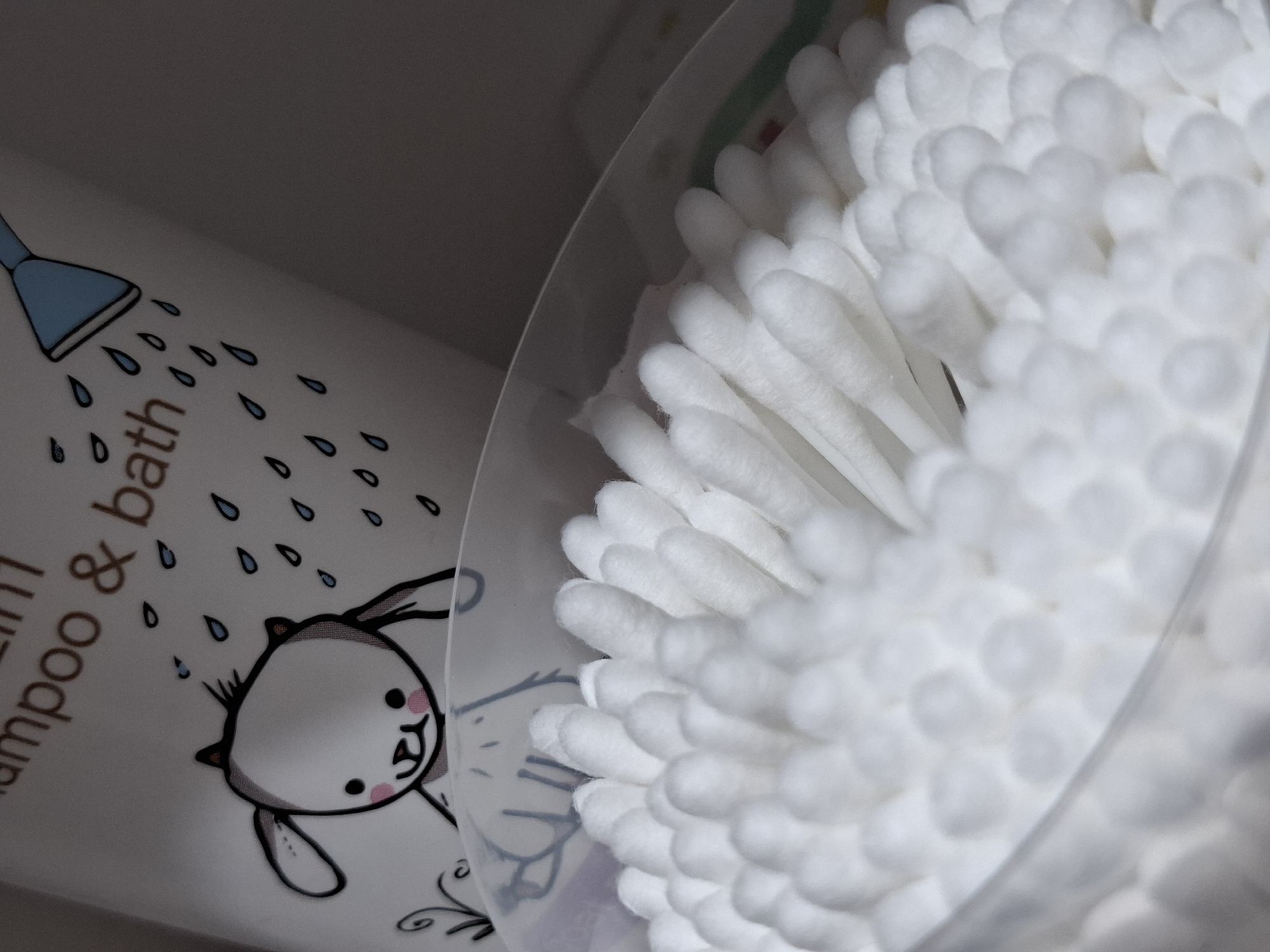 This close-up shot features two personal care items, likely situated in a bathroom or nursery setting. On the right, a large number of white cotton swabs are neatly arranged upright within a transparent, cylindrical container. The cotton tips, soft and fluffy, fill the container, creating a textured pattern. The plastic container's rim is visible, subtly separating the cotton swabs from the other item.
To the left, a portion of a white or light-colored bottle is visible, adorned with cheerful illustrations and text. A cartoon bunny, rendered in black outline with pink cheeks and ear details, is depicted looking upwards. Above the bunny, a blue shower head with a black outline is shown emitting numerous blue water droplets, some of which appear to be splashing onto the bunny. Partial text is visible on the bottle; the words "shampoo & bath" are clearly discernible, with "in 1" also visible, suggesting a "2-in-1" or similar product. A word ending in "ath" (likely completing "bath") is also present. The overall impression is of baby or child-friendly bath products. The items are illuminated, creating soft shadows and highlights that emphasize their textures and shapes.