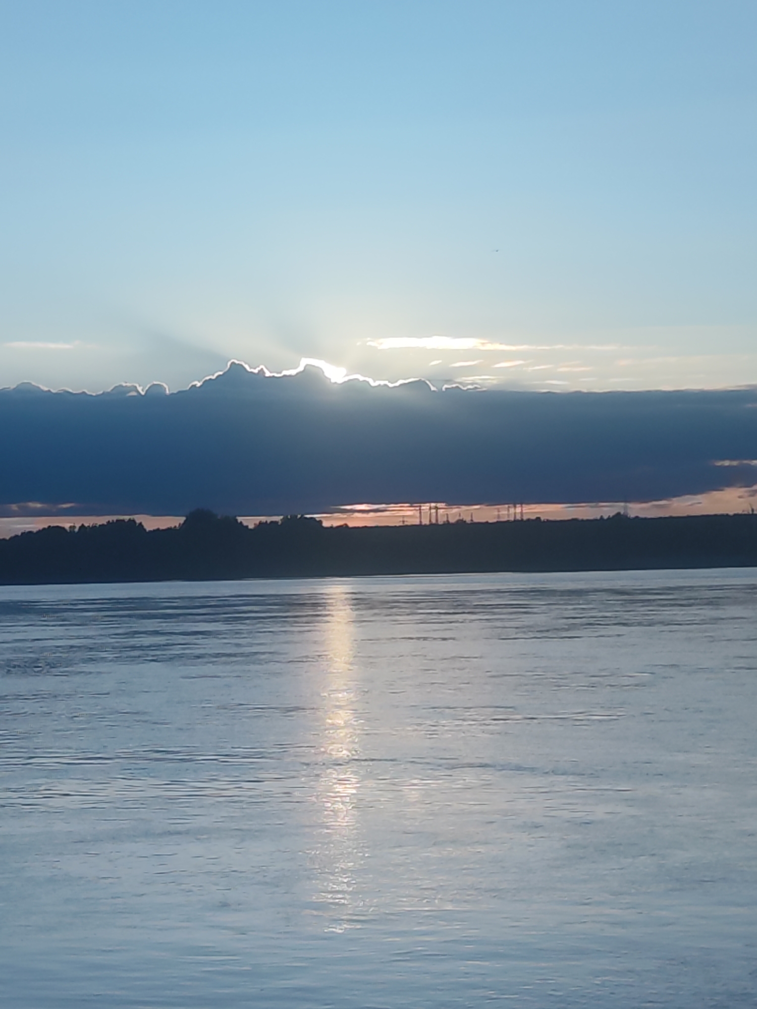 The image captures a serene landscape featuring a vast body of water under a dramatic sky, likely at sunrise or sunset. The sky transitions from a clear light blue at the top to warmer hues near the horizon, where the sun is partially obscured by a thick, dark cloud bank. Bright sunbeams (crepuscular rays) pierce through gaps in the clouds, creating striking shafts of light that extend upwards.
A distant landmass forms the horizon, silhouetted against the sky. This distant shore appears to be covered in trees, with some industrial structures, possibly power lines or communication towers, faintly visible above the treeline.
The calm surface of the water reflects the sky and the sun's light, creating a shimmering, elongated path of brightness extending from the horizon towards the foreground. Small ripples are visible on the water's surface. A tiny, dark silhouette of an aircraft, possibly a plane or drone, can be seen high in the clear part of the sky, above the main cloud formation. The overall impression is one of tranquility and natural beauty, highlighting the interplay of light, water, and clouds during a golden hour. No visible text is present in the image.