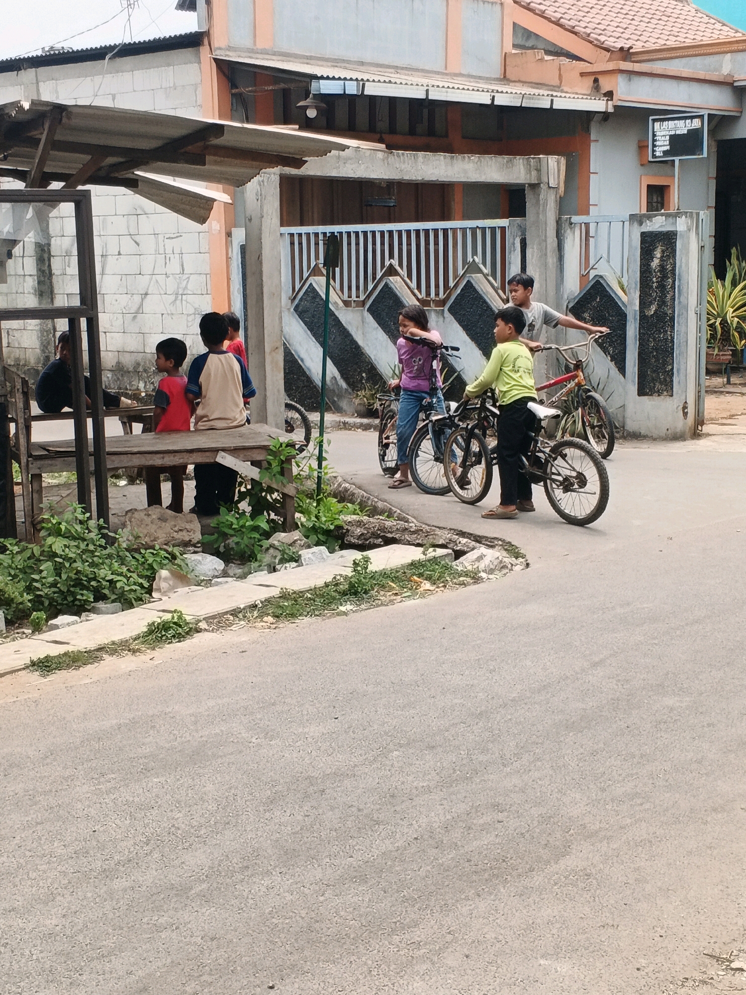 The image shows a street scene in Kadu, Indonesia, with several children and bicycles. In the foreground, a gray asphalt road curves gently. To the left, a wooden bench structure is partially covered by a corrugated metal roof supported by wooden posts. Three young boys are seated on the bench, facing right. The boy on the far left wears a black t-shirt and appears to be facing away from the camera. The middle boy has short black hair and wears a red t-shirt and blue pants. The boy on the far right has short black hair and wears a light brown long-sleeved shirt with a blue collar and dark pants.
Further back, several children are interacting with bicycles. A girl in a purple shirt and blue jeans stands on the left, holding the handlebars of a pink bicycle. To her right, a boy wearing a light green long-sleeved shirt and dark pants stands with his hands on the handlebars of a black bicycle. Behind them, another boy in a gray t-shirt leans on the handlebars of a red bicycle. A man in a gray t-shirt stands behind this boy, his hands also on the handlebars.
In the background, a building with a light brown tiled roof and light gray walls is visible. A fence with black and white diamond patterns runs along the front of the building. A black sign with white text is mounted on the building's wall. Green foliage is present along the side of the road and near the building. The overall scene suggests a casual gathering of children in a residential or semi-public area.