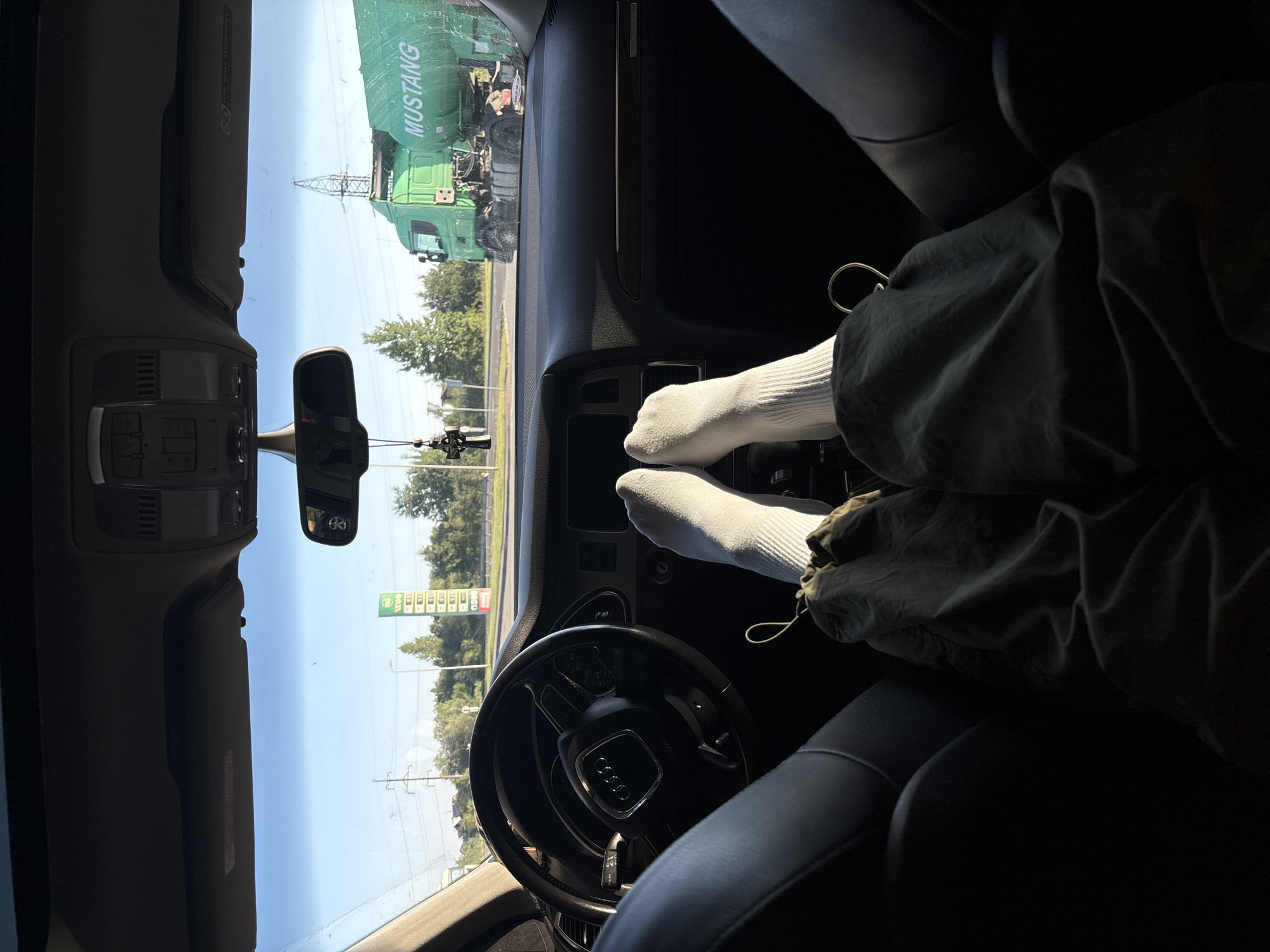 The image is taken from the interior of a car, looking forward through the windshield. The perspective is from the passenger seat, with the viewer's feet and legs, clad in white socks and dark pants, visible in the foreground. The dashboard and steering wheel of an Audi are in the lower center and left of the frame.
Outside the car, the scene appears to be a road with a gas station to the left and a large green tanker truck to the right. The truck has "MUSTANG" written on its side. The sky is clear and blue, suggesting it is daytime. The environment seems to be suburban or on the outskirts of a town, with trees and power lines visible in the background.
The image is not specific to the city of Tysmenytsia, Ukraine, as there are no identifiable landmarks or text that would confirm this location.