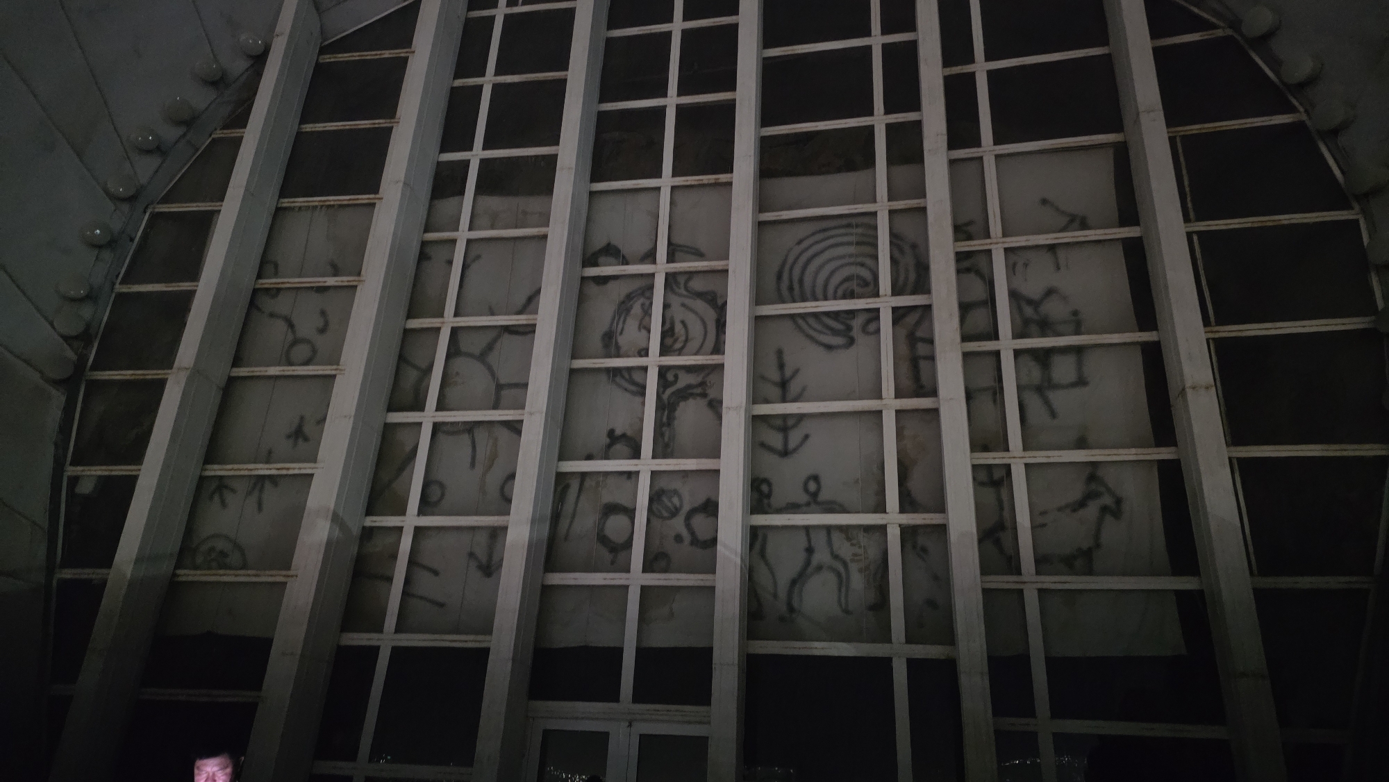 The image shows a close-up of a large, curved window or facade made of many rectangular panes divided by white metal framing. Behind the framing, a large piece of light-colored fabric or sheeting is visible, on which numerous dark, abstract drawings or symbols are spray-painted. These markings appear primitive, with some resembling stick figures, spirals, and geometric shapes. The overall scene is dimly lit, with a portion of a person's face, partially obscured by shadow, visible in the lower left corner of the image. The architecture suggests an indoor space, possibly a hall or a large building with a distinctive curved design, located in Osh City, Kyrgyzstan, based on external information.