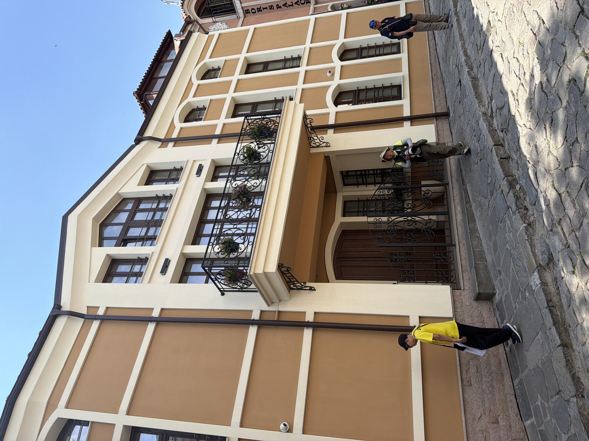 The image depicts a detailed view of a cobblestone street and an ornate building under a clear blue sky in Plovdiv, Bulgaria.
The central and dominant structure is a multi-story building featuring a warm brown facade with cream-colored trim outlining windows, corners, and architectural details. The building has dark brown window frames and a prominent central double wooden door with an intricate wrought-iron gate in front. Above the entrance, a balcony with a black wrought-iron railing, decorated with potted plants, extends outwards, supported by ornate black metal brackets. A dark brown drainpipe runs vertically along the left side of the facade. A white security camera is mounted on the left wall section at a lower level. The building's base is clad in rough-cut, light-colored stone. To the right, a section of an adjacent building is visible, featuring a vertical sign that reads "BORIS PALACE" and a red-tiled roof.
Three men are present on the cobblestone street. On the far left, a man walks from right to left, wearing a black baseball cap, a bright yellow t-shirt, black pants, and dark athletic shoes. He carries a white cloth or bag and a black shoulder bag. In the center, a second man walks from left to right, wearing a light-colored sun hat, a light-colored long-sleeved shirt with a dark vest or straps, olive green pants, and light-colored shoes. He holds a large camera with a telephoto lens and has a small black bag attached to his belt. On the far right, a third man stands, facing slightly towards the viewer, wearing a blue baseball cap, a dark blue shirt, light brown pants, and light blue athletic shoes. He carries a black shoulder bag.
The street is paved with irregular grey cobblestones, showing some minor vegetation growth in the cracks. Strong shadows from the buildings and people are cast onto the street, indicating bright daylight conditions. A faint, small object is visible in the upper right section of the clear blue sky.
