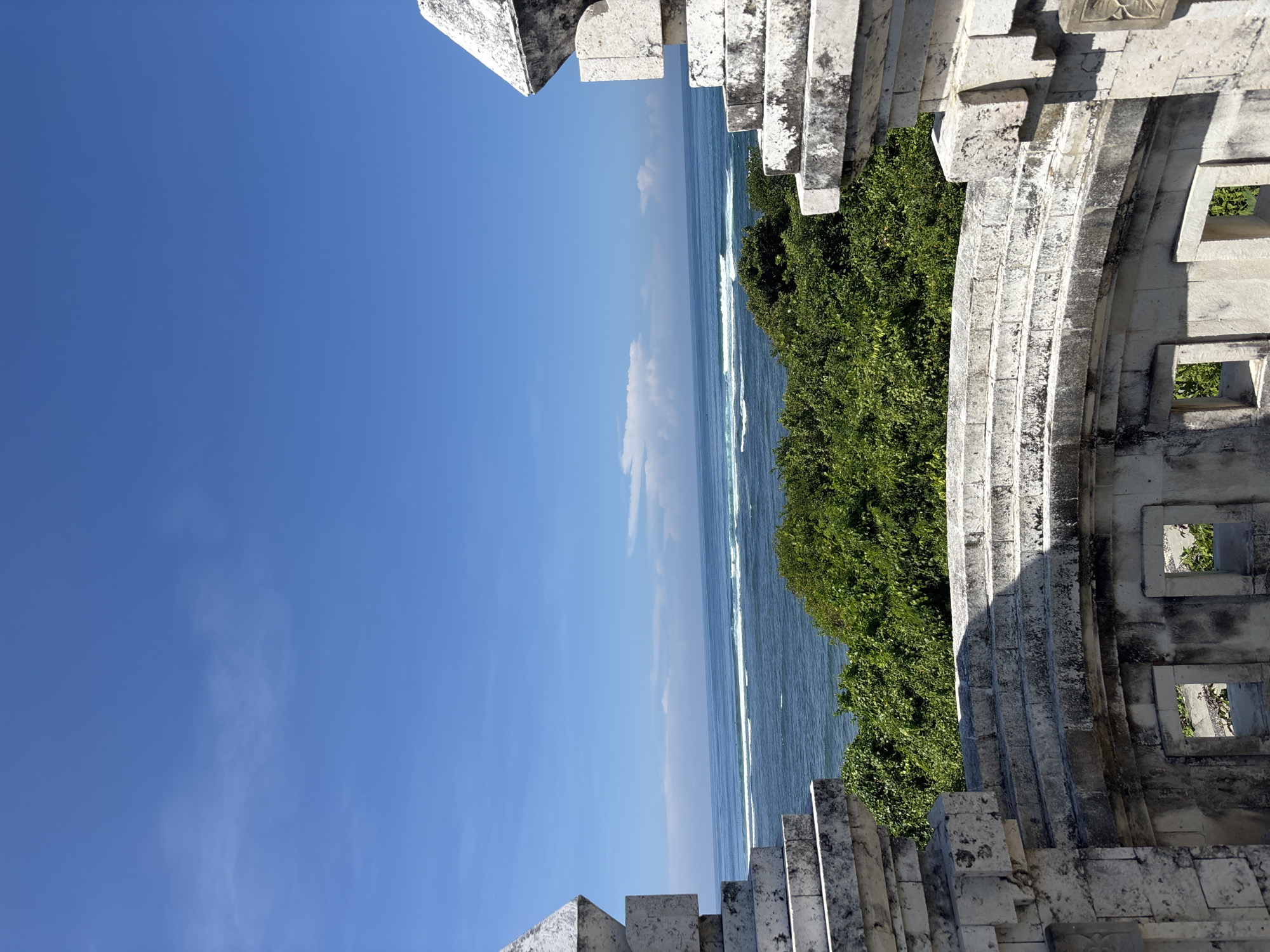 The image shows a view of the ocean and sky from what appears to be the Uluwatu temple in Kuta Selatan, Bali, Indonesia. The framing of the image is provided by the temple's stonework architecture on both sides. Vegetation is seen between the stonework and the ocean. The ocean is pale blue with white wave crests. The sky is a clear, bright blue with some wisps of clouds.