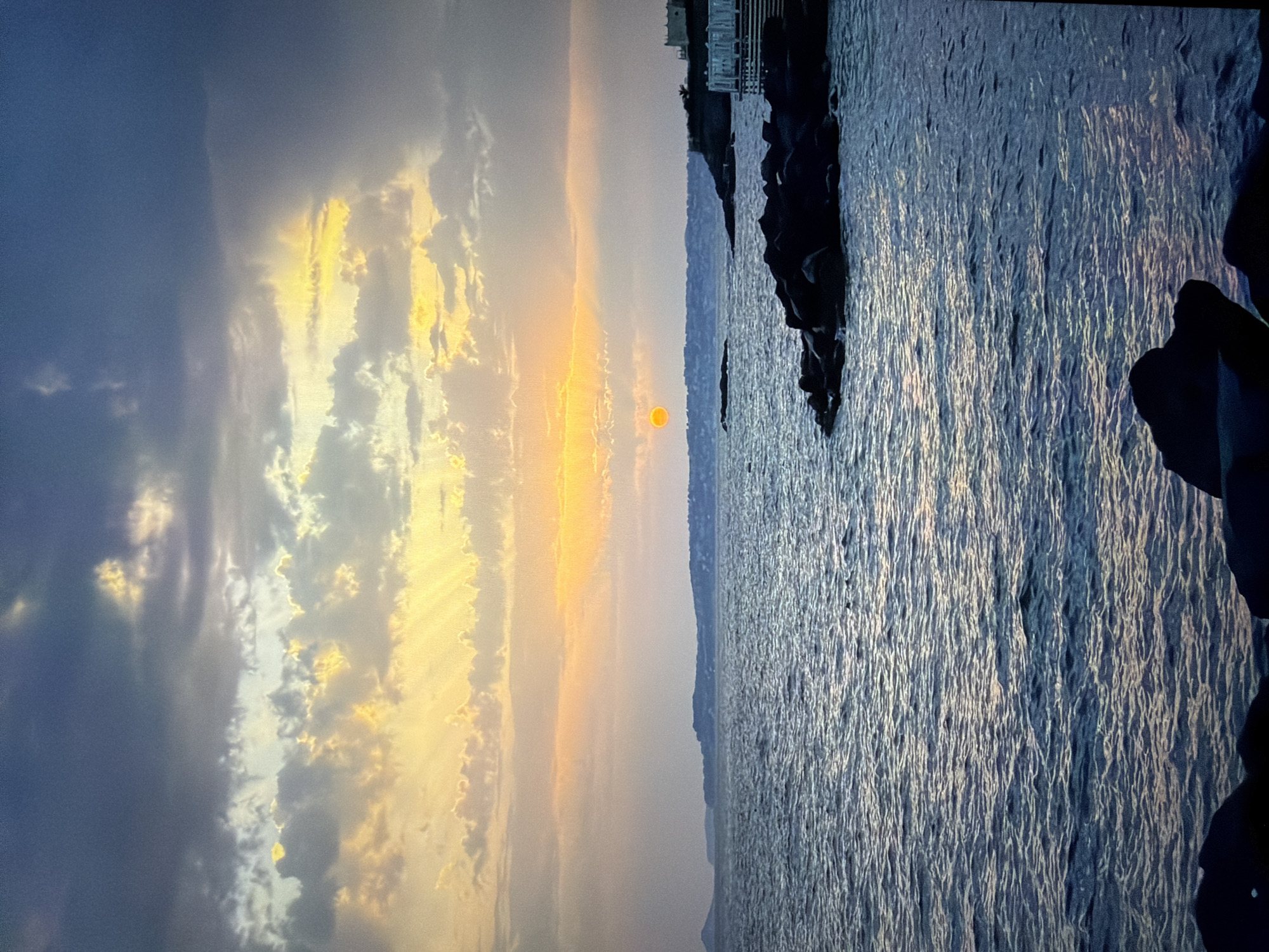 This media file captures a serene coastal sunset scene at Ros Na Rí, Ireland. The image, presented in a portrait orientation but depicting a horizontal landscape, showcases a dramatic sky and a vast body of water.
The sky, filling the upper half of the frame, displays a striking contrast of colors. On the far left, dark grey-blue clouds dominate, gradually transitioning towards the center and right into brilliant golden-yellow and orange hues. These clouds are heavily illuminated by the setting sun, creating vivid patterns of light. The sun itself is a distinct, bright orange orb, positioned low on the horizon line, just above a distant landmass.
The lower half of the image features a large body of water, likely the sea, with a textured, deep blue-grey surface. It reflects the vibrant sky in shimmering streaks, particularly towards the central and right portions, suggesting light ripples or waves. Along the right side of the frame, dark, jagged rocky formations or a breakwater extend into the water, appearing as stark silhouettes against the lighter backdrop.
A faint, hazy landmass or distant coastline stretches across the horizon beneath the sun. Above the dark rocks on the far right, a subtle outline of a building or structure is vaguely visible on this distant shore. The scene is devoid of people or visible text, conveying a sense of tranquil natural beauty during dusk.