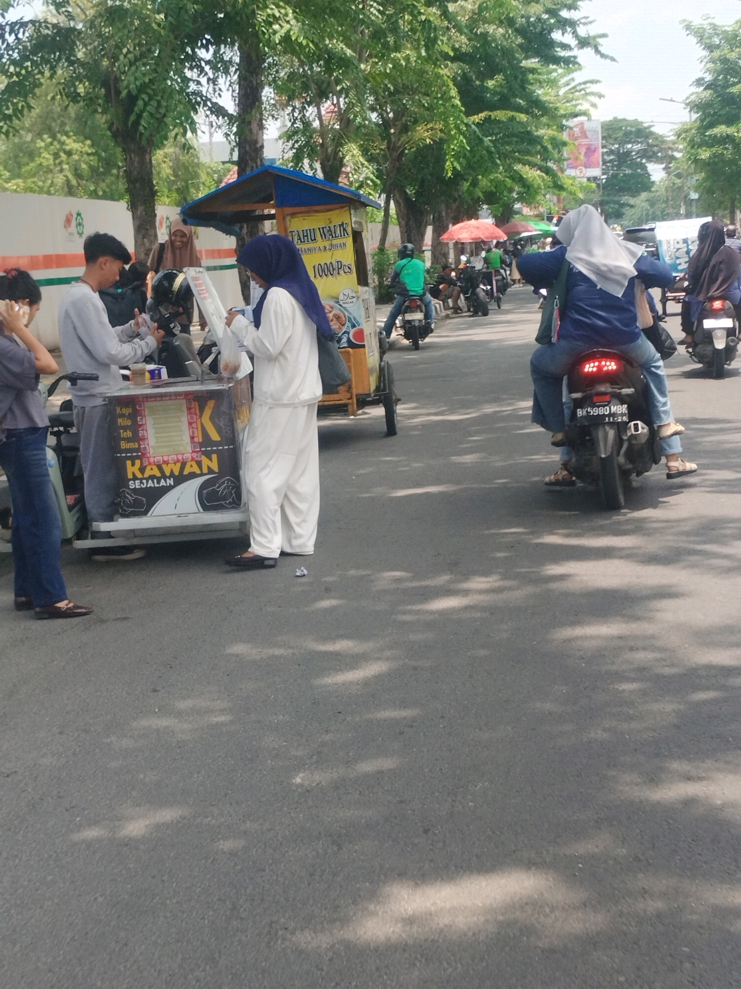 In the city of Medan, Indonesia, a street scene unfolds with a focus on a small food cart. The cart, adorned with the text "Tahu Walik," "Hanya & Jihad," and "1000 Pcs," is positioned on the side of a paved road. A vendor, a woman wearing a hijab and white clothing, stands behind the cart, interacting with customers. Another person, also wearing a hijab, is observing the transaction. In the foreground, a young woman in a gray shirt and blue jeans stands on the left, looking towards the cart. Behind her, a young man in a gray long-sleeved shirt and dark pants is engaged with the vendor, holding what appears to be a receipt. Two men on motorcycles are also visible. One is wearing a green shirt and a black helmet, and the other is wearing a dark jacket and helmet. In the background, several other individuals on motorcycles are present, and trees with green foliage line the street. The overall environment suggests a typical urban street in Indonesia.