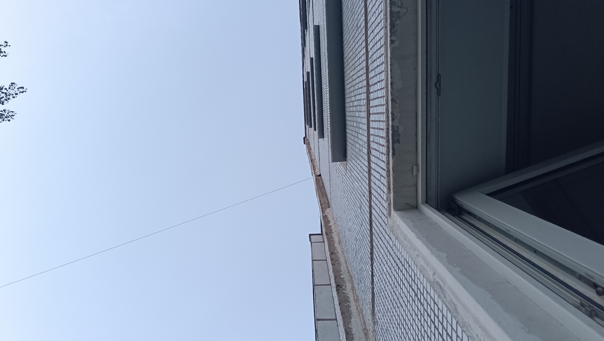 The image shows a low-angle view of the exterior of a tiled building, possibly an apartment building, under a clear, pale blue sky. The building has several horizontal windows with light-colored frames, some of which appear to be open, revealing dark interiors. A thin, dark line, possibly a wire or string, stretches diagonally across the sky from the left side of the frame towards the building. On the far left, the dark silhouette of tree branches is visible against the sky. The building's facade is covered in small, square, light-colored tiles, with some areas showing wear or discoloration, particularly around the window frames. The lighting suggests it is daytime. The scene is set in the city of Chaikovsky, Russia.