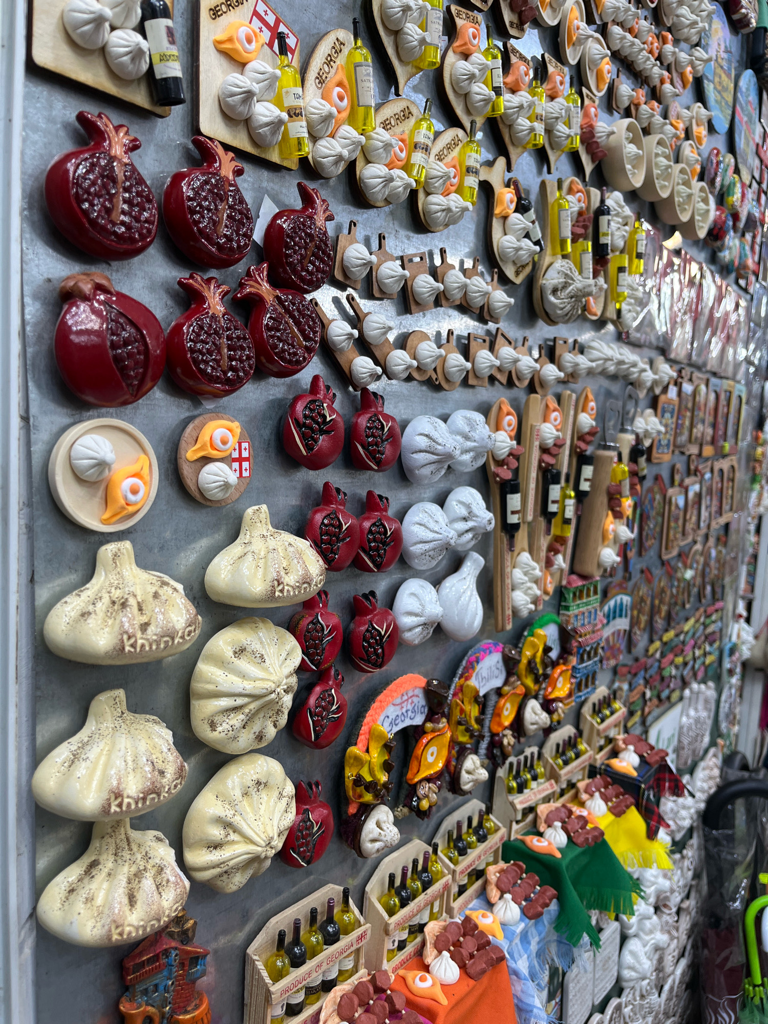 The image shows a close-up view of a wall covered with souvenirs in Tbilisi, Georgia. The souvenirs are primarily magnets, depicting various Georgian cultural items and food. Prominently featured are magnets shaped like pomegranates, khinkali (dumplings), bottles of wine, and traditional Georgian flags. There are also magnets representing khachapuri (cheese bread) and skewers of meat. The souvenirs are arranged in a dense, somewhat disorganized manner, covering a significant portion of the wall's surface. The overall impression is of a bustling market or shop selling local crafts.