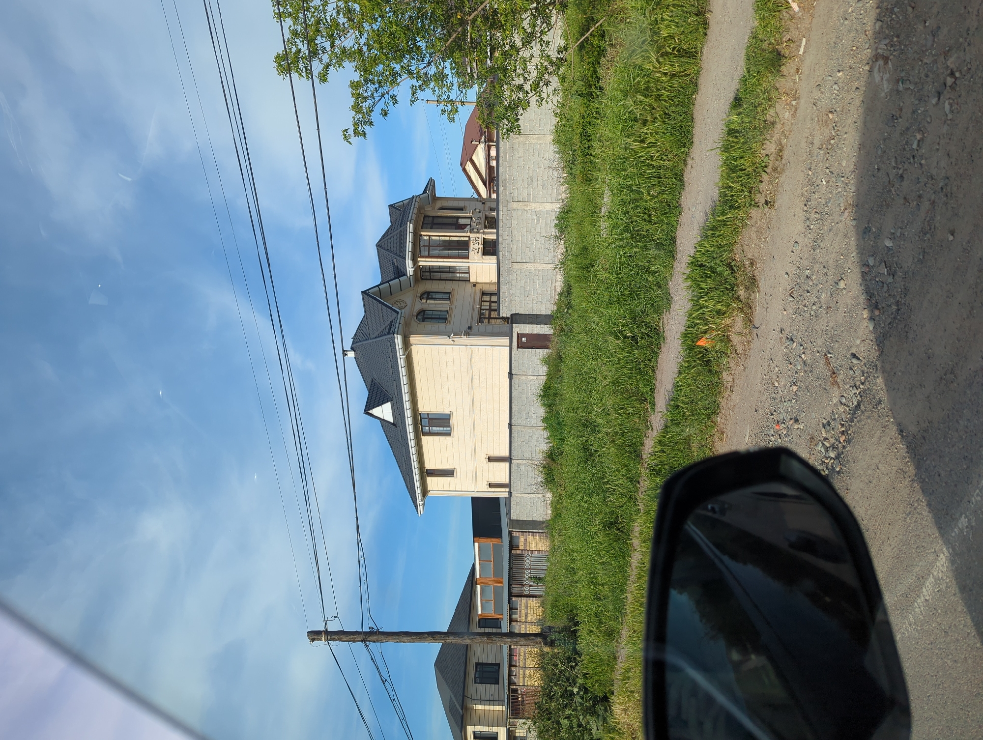 A three-story beige house with a dark gray roof is adjacent to a gravel road in Beş-Küŋgöy, Kyrgyzstan. The house is surrounded by a concrete block fence and green grass. A wooden utility pole with power lines is visible in the foreground. A car side mirror is partially visible in the lower right corner. The sky is blue with white clouds. Another house is visible in the background behind the primary house.