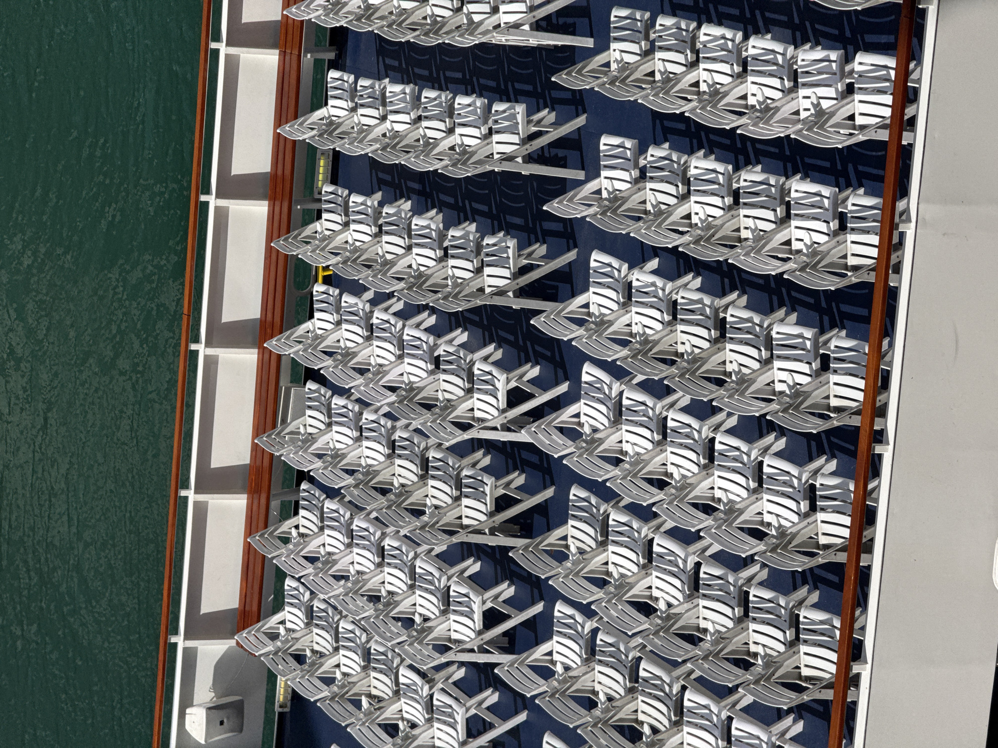 The image is an overhead view of rows of white folding chairs stacked neatly on the deck of a ship, with dark blue carpeting beneath. A wooden railing runs along the edge of the deck, separating it from a dark green body of water. The chairs are arranged in a grid-like pattern, with their backs facing the viewer. The lighting suggests a sunny day, with distinct shadows cast by the chairs. There are no people visible in the image. This scene is not located in Chicago, United States of America.