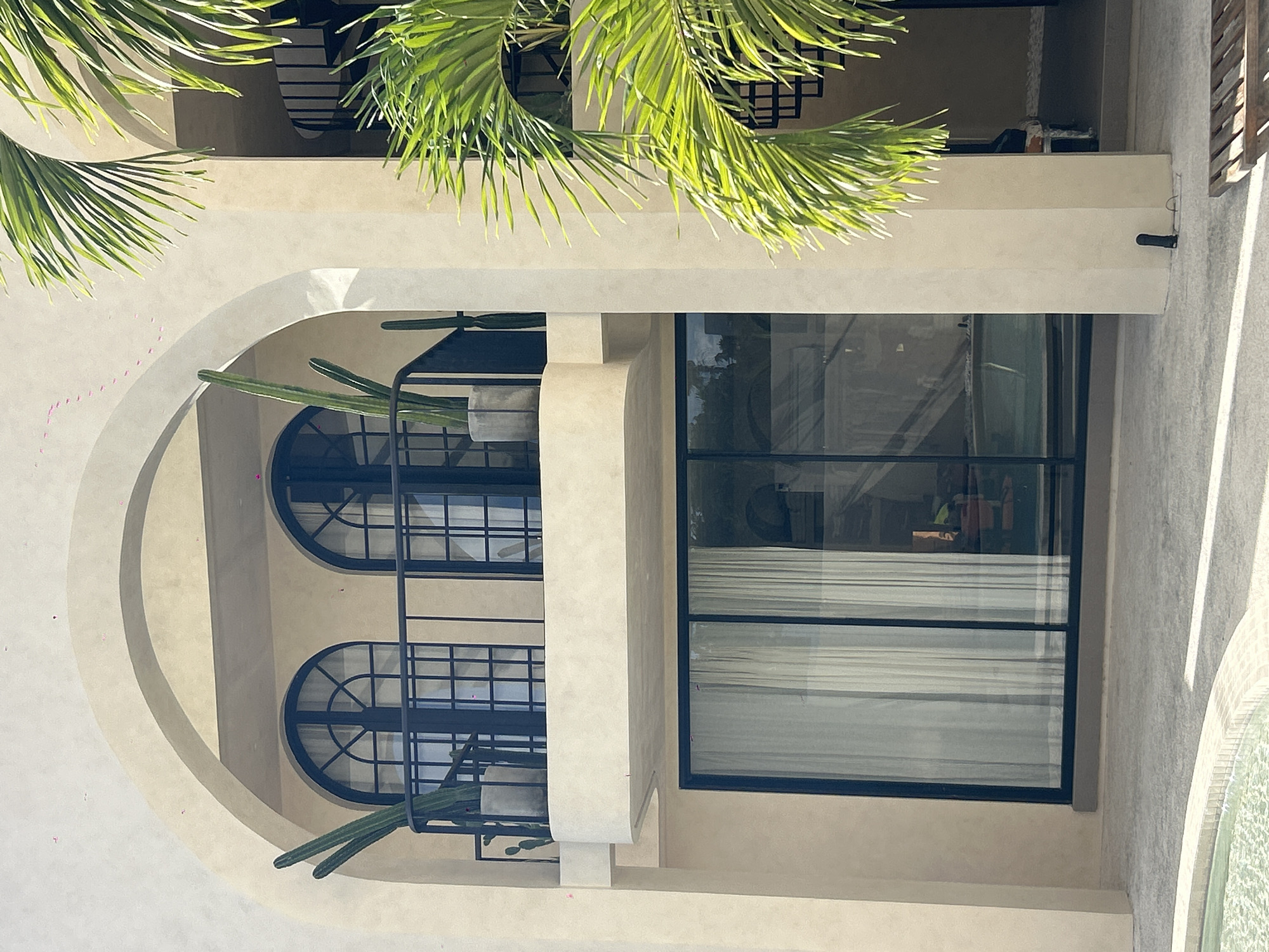 The image shows a modern, stucco building with arched openings and balconies. Two large arched windows with dark frames and grid patterns are visible on the upper level, with a black metal railing in front of them. Several tall, green columnar cacti are potted on the balcony. Below, a large, floor-to-ceiling window with a dark frame spans the width of the building. Sheer white curtains are drawn behind the glass. To the right of the building, a portion of a spiral staircase and vibrant green palm fronds are visible. The ground in front of the building is paved, and a glimpse of a tiled pool is visible in the lower left corner. The architecture suggests a tropical or Mediterranean influence. This scene is located in North Kuta, Indonesia, a region known for its resorts and coastal attractions. There are no people visible in the image.