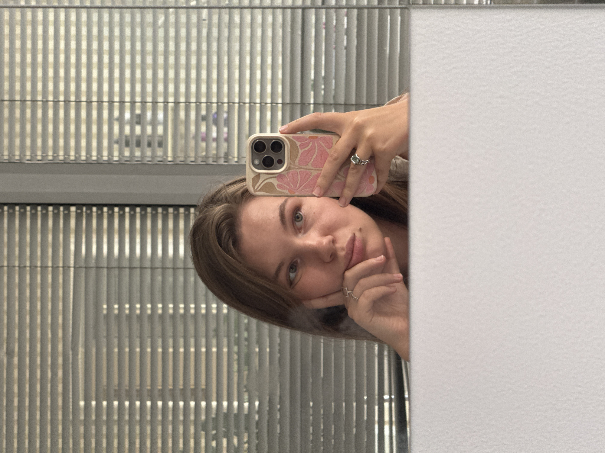 A young woman is taking a mirror selfie in what appears to be a residential setting. She has light brown hair, fair skin, and is wearing two silver rings. She is holding a smartphone with a pink floral case in her right hand. Her left hand is resting on her chin. The background consists of horizontal blinds, suggesting a window. The foreground shows a textured white surface, possibly a countertop or furniture. There are no discernible city landmarks or geographical indicators present to identify the location within Vracar Urban Municipality, Serbia.