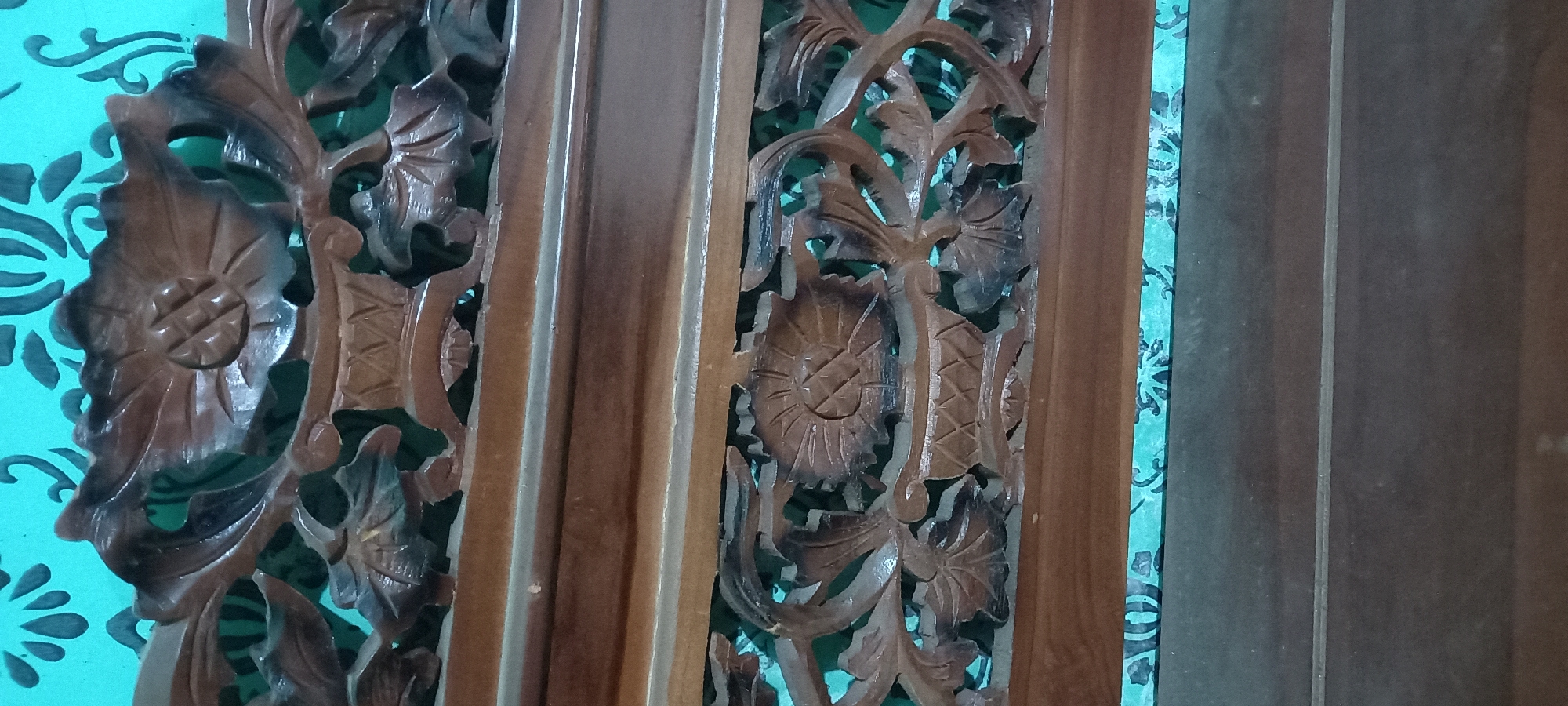 The image shows a close-up of intricately carved wooden furniture, likely a part of a bench or a decorative panel. The wood is dark brown with a glossy finish, highlighting the detailed floral and basket-like motifs. Behind the carving, a vibrant turquoise background with a dark, repeating floral pattern is visible. The overall impression is one of traditional craftsmanship and artistry. The location specified as Gulurejo, Indonesia, suggests this is an example of Indonesian woodworking or decor.