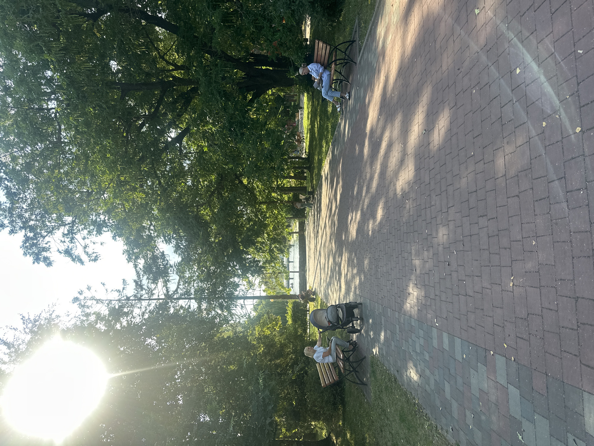 The image depicts a sunlit park path in Ternopil, Ukraine. The path, paved with reddish-brown and gray interlocking bricks, is lined by lush green trees casting dappled shadows.
On the left side of the path, a woman with blonde hair, wearing a white t-shirt and light-colored pants, sits on a wooden park bench. Next to her is a dark gray baby stroller. Further down the path, in the distance, another person is visible sitting on a bench.
On the right side of the path, a woman with blonde hair, wearing a light blue long-sleeved shirt and blue jeans, sits on a wooden park bench, reading a book.
In the background, a body of water, possibly a lake or river, is visible, reflecting the sunlight. A bridge or structure can be seen crossing the water in the distance. The overall atmosphere is peaceful and suggests a pleasant day in the park.