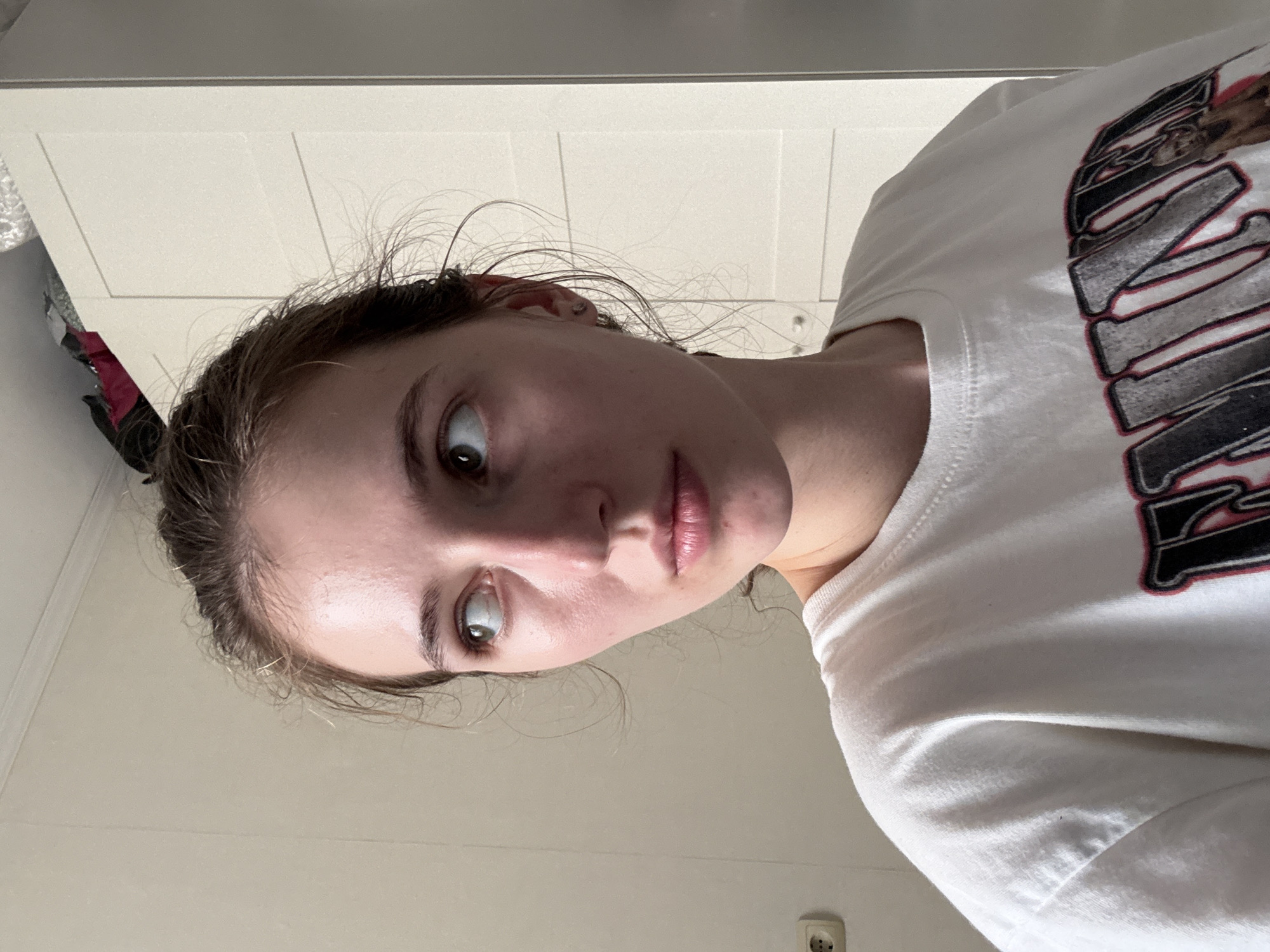 A young woman with light brown hair pulled back in a bun is looking to her left. She is wearing a white t-shirt with a graphic design on it. The background is a plain wall with a light switch and an outlet on the left side. To the right of the woman is a white wardrobe. There are items on top of the wardrobe, some of which appear to be bags. The image appears to be taken indoors in a domestic setting. There is no information in the image to suggest the location is the city of Perm, Russia.