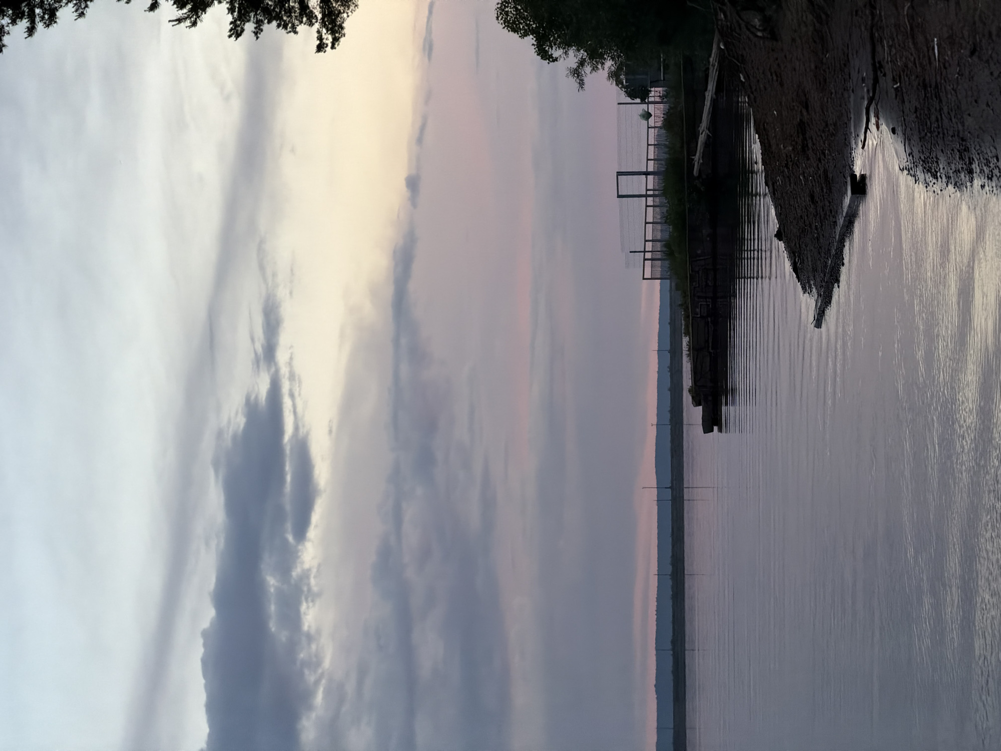 The image shows a body of water reflecting a cloudy sky at dusk. A shoreline with vegetation and a partially visible fence runs along one side of the frame. Tall, slender structures rise from the water near the shoreline. The image is taken outdoors and depicts a serene scene. It is difficult to pinpoint the precise location within Chaikovsky without further information.