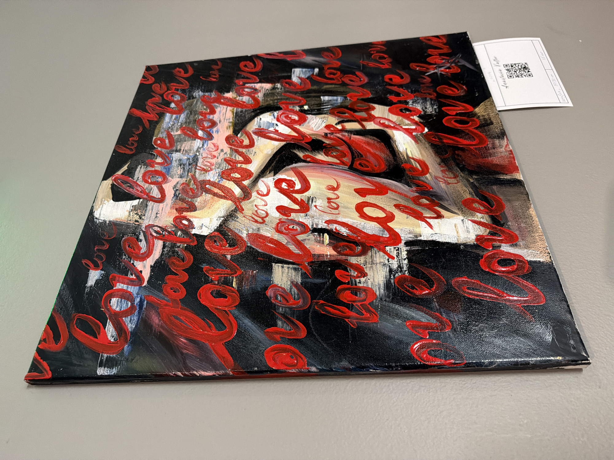 The image depicts an abstract painting on a square canvas. The artwork features a nude figure partially obscured by the word "love" repeatedly written in bold, red brushstrokes. The background is primarily black with streaks of white, tan, and blue. A small tag with a QR code and the name "Anastasia Valtor" is attached to the top right corner of the canvas. The canvas is resting on a light gray surface. The location is Unknown, Unknown.