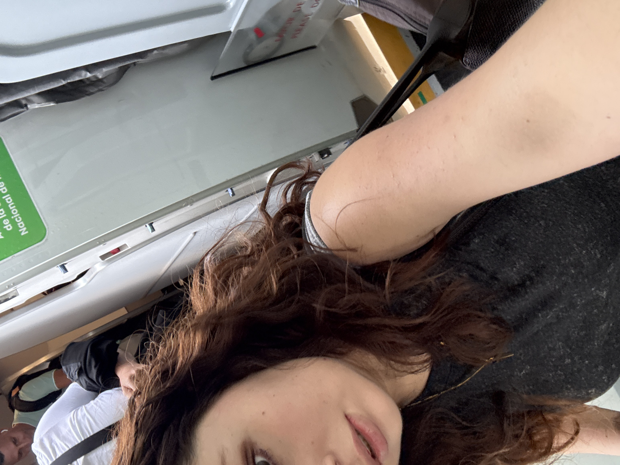 A selfie-style image captures a woman from a low angle within what appears to be a vehicle or public waiting area in Mexico City, Mexico. The main subject, a woman with long, curly, dark brown hair, looks directly at the camera with a neutral to slightly smiling expression. She wears a dark grey or black top and a thin gold chain necklace. Her fair-skinned arm is prominently featured in the foreground, showing minor skin texture and a few small moles.
The background reveals the interior of the setting, characterized by large, white, possibly plastic or metal panels, suggesting a functional, institutional, or transport environment. A bright green sign with white text is visible, partially reading "Nacional de la" and "De la." Further in the background, some red text on a lighter surface can be discerned, including "PLEASE DO" and a word resembling "PRECAUCIÓN" (caution) in Spanish, reinforcing the public or regulated nature of the location. Other individuals are partially visible in the background, appearing to be seated or standing. The lighting is bright and even, indicating daytime. The overall impression is that the woman is taking a photo while traveling or waiting in transit.