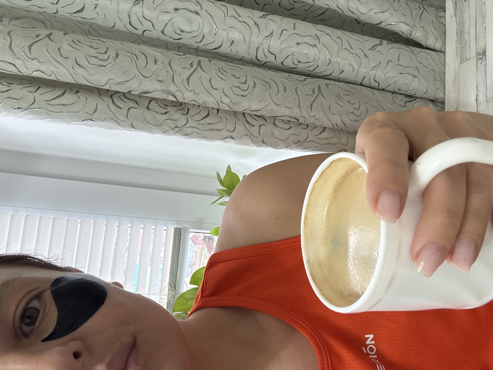 The image is a close-up selfie taken indoors. A person with fair skin and dark hair is visible, wearing a black eye patch under their left eye and an orange tank top. They are holding a white mug filled with a light brown liquid, likely coffee or tea. The background includes white blinds, a potted plant, and decorative curtains with a swirling pattern. While location details are not available in the image, the user has requested for it to be in the city of Chaikovsky, Russia.