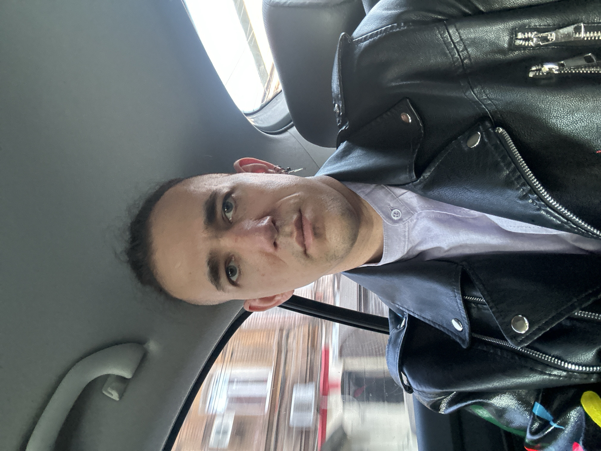 A person is seated in what appears to be a car, visible through the window is buildings characteristic of the city of Saratov, Russia. The person is wearing a black leather jacket over a light purple button-down shirt. The car's interior is gray. The image's perspective is from a slightly upward angle relative to the person.