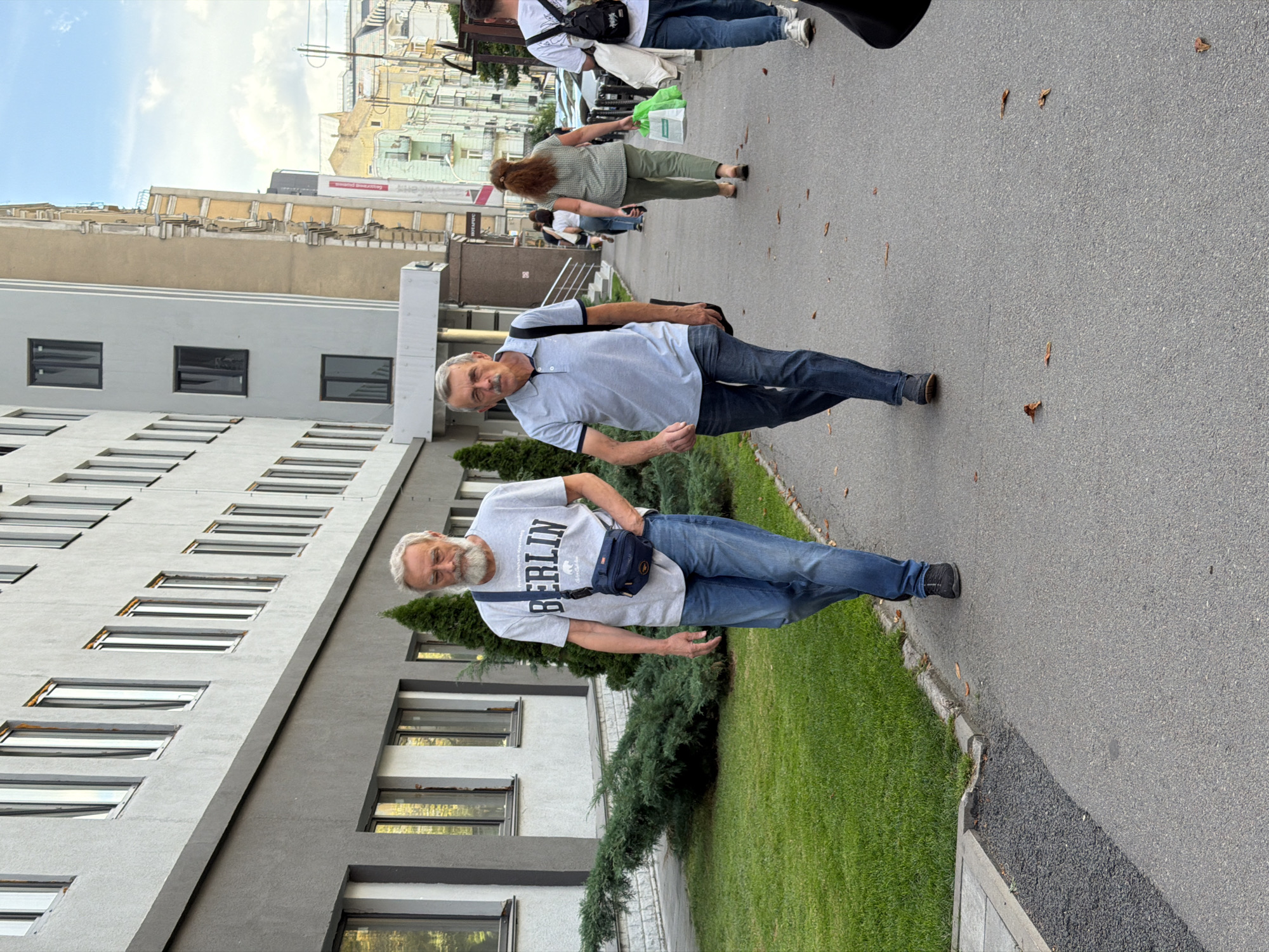 Two men are walking on a sidewalk in front of a modern building. The man on the left is older with a grey beard and is wearing a grey t-shirt with "BERLIN" printed on it, blue jeans, and a navy blue fanny pack. The man on the right is also older, with a mustache, and is wearing a light blue polo shirt, blue jeans, and carrying a dark bag slung over his shoulder. The building behind them is light grey with numerous windows. Green grass and bushes are visible to the left of the sidewalk. In the background, other pedestrians and buildings can be seen. The sky is partly cloudy. The scene is captured outdoors in daylight.