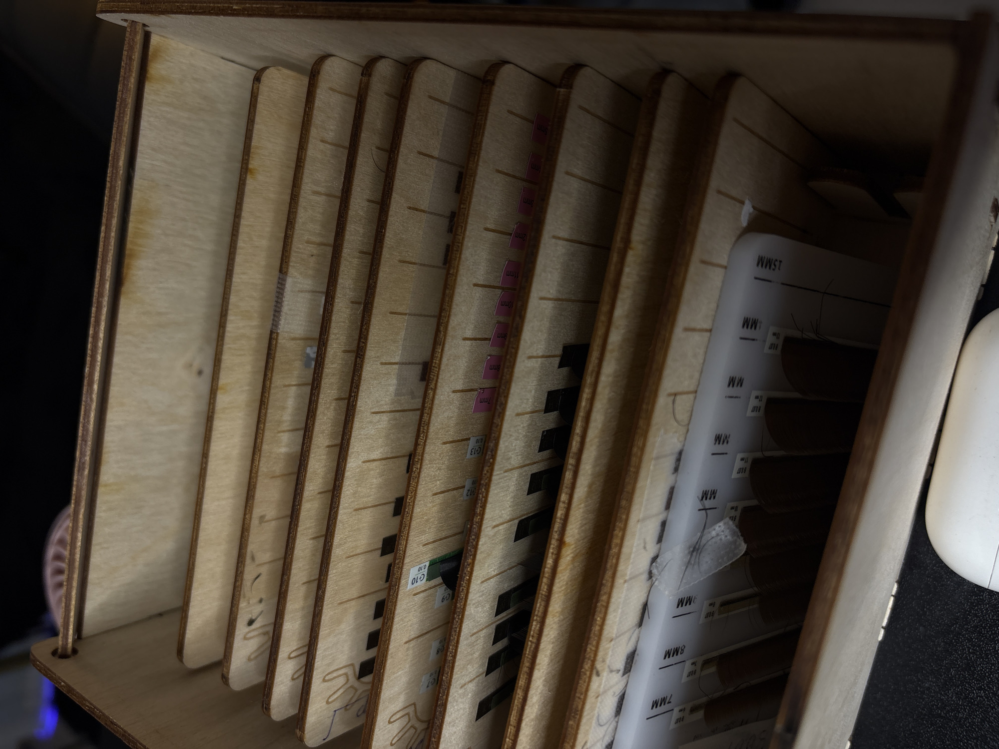 This media file captures a close-up, eye-level view of a wooden organizer filled with trays of false eyelashes. The organizer is made of light-colored wood with visible grain and appears to be a storage solution for beauty supplies. Each tray is compartmentalized, and the false eyelashes are arranged in neat rows, varying in length.
Several trays have labels indicating different sizes, such as "7MM," "8MM," "9mm," "10mm," "11mm," "12mm," and "15MM." Some labels are pink with black text, while others are white with black text. Some trays also display codes like "C13," "C12," "C10," "C9," and "C7," along with numbers like "0.10." A small piece of clear tape with a few stray eyelashes is attached to one of the trays. The lighting suggests an indoor setting, possibly a beauty salon or a personal makeup station, with the focus on the organized collection of eyelashes. The scene is static, with no discernible activity taking place other than the storage of these items. The overall impression is one of order and preparedness in a beauty context. The scene lacks any human presence, weather cues, or specific time of day indicators.