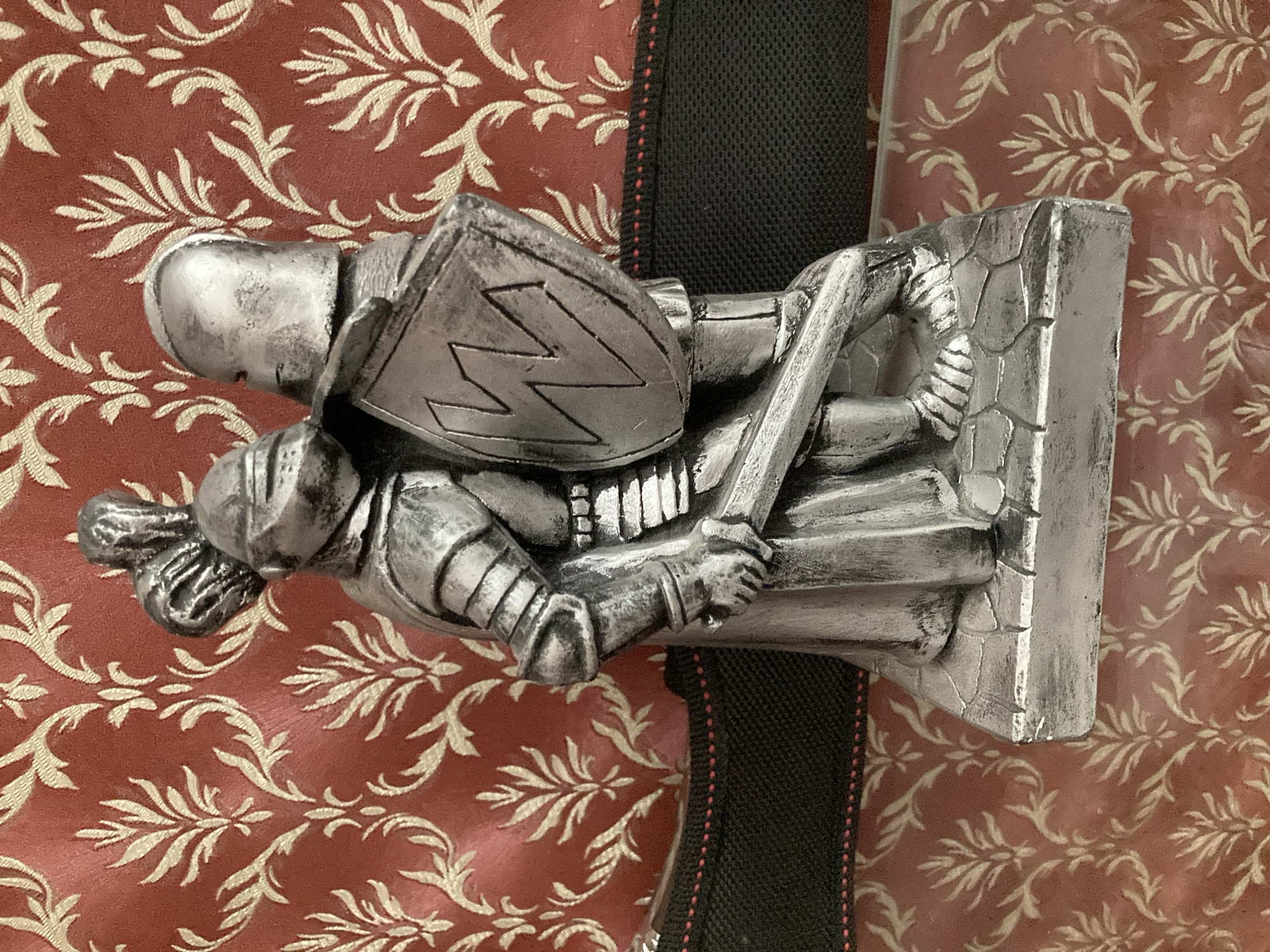 A silver-colored statue of two knights in armor stands on a grey base in front of a red damask patterned background. The knight on the left is slightly taller and wears a helmet with a plume. He holds a sword in his right hand and a shield in his left. The knight on the right is shorter and wears a helmet with a visor. He is facing the other knight and appears to be holding the shield with the left knight. The statue is positioned on a surface that is covered by the damask pattern. There is a black fabric band with red stitching visible behind the statue. The scene is lit from above, casting shadows. The location is Unknown, Unknown.