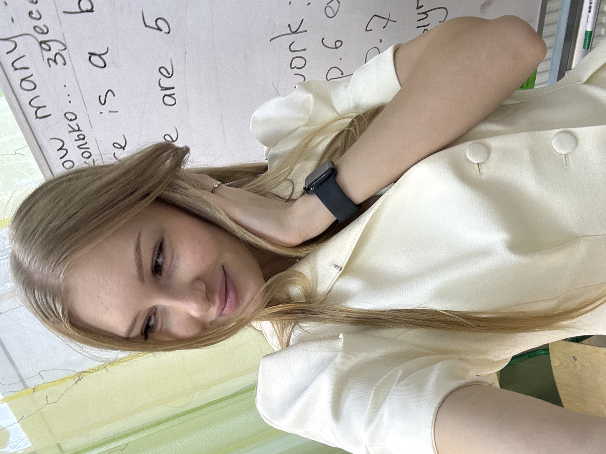 A young woman with blonde hair is captured in a medium shot, facing slightly to her left with her head tilted back. She is wearing a cream-colored jacket with puffed sleeves and is touching her hair with her right hand. On her left wrist, she wears a dark smartwatch. The background features a whiteboard with handwritten text in English and Russian. The text includes phrases like "How many," "сколько ...," "there is a," and "are 5." There are also other handwritten notations and markings on the board. The environment appears to be an indoor setting, possibly a classroom or office, with a green curtain or blind visible on the left side of the frame. The overall lighting is bright, highlighting the woman and the whiteboard. The image quality is clear, showing details of the woman's facial features and the textures of her clothing.